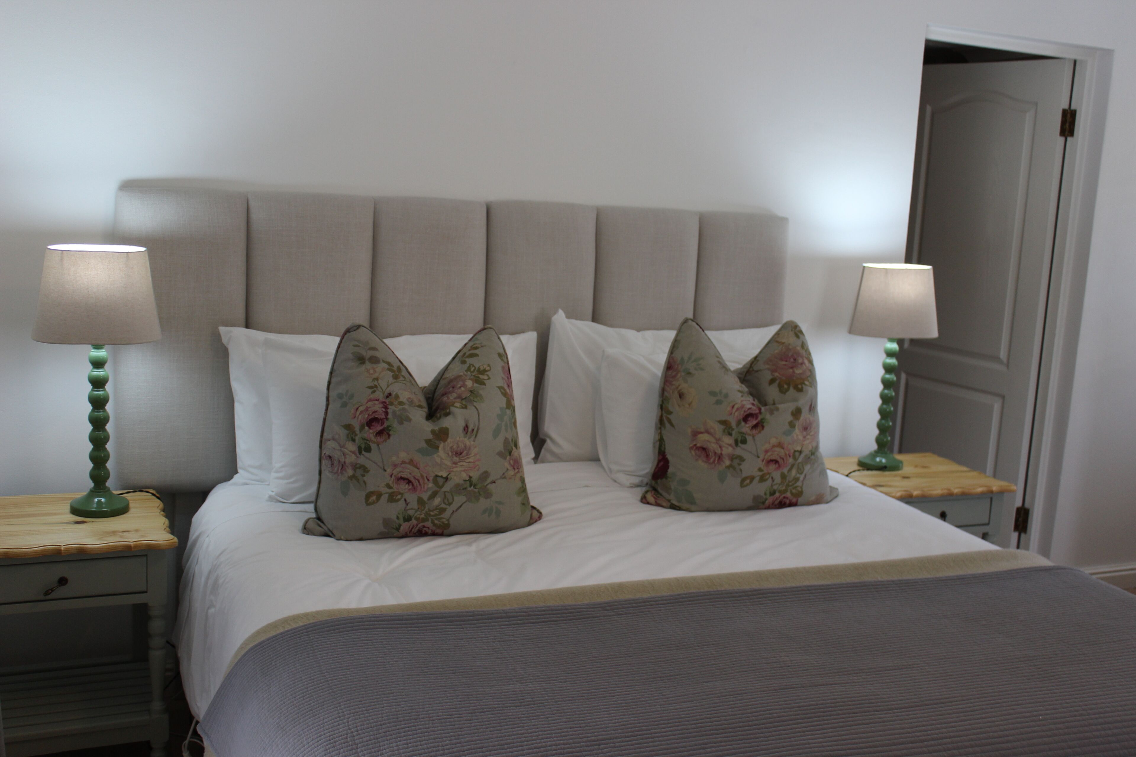 Cama bem arrumada com almofadas florais, duas luminárias de mesa e uma mesinha de cabeceira de madeira.