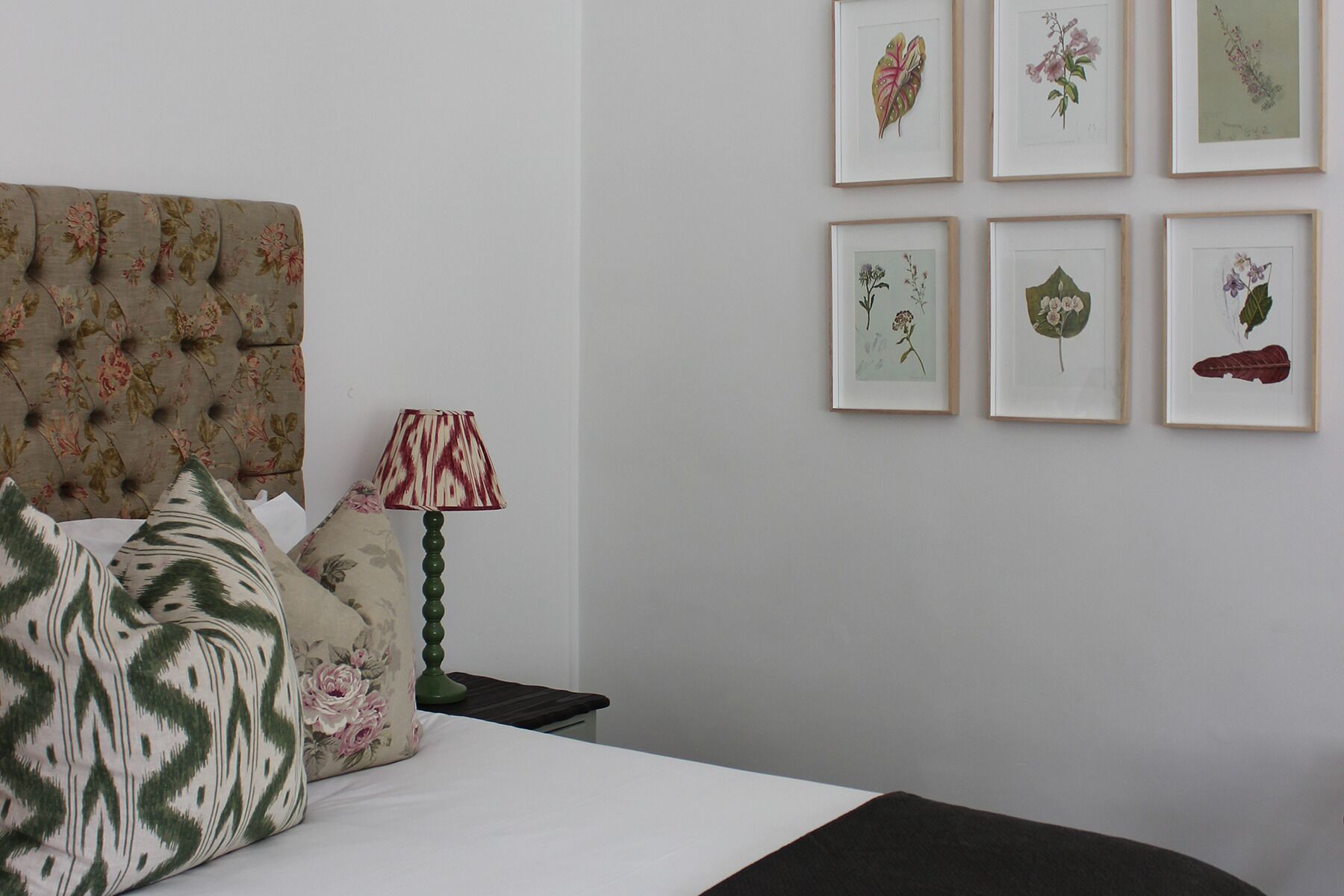 A bedroom with a tufted headboard, floral-patterned pillows, a bedside lamp, and framed botanical prints on the wall.
