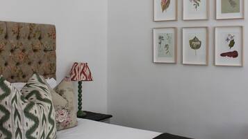 A bedroom with a tufted headboard, floral-patterned pillows, a bedside lamp, and framed botanical prints on the wall.