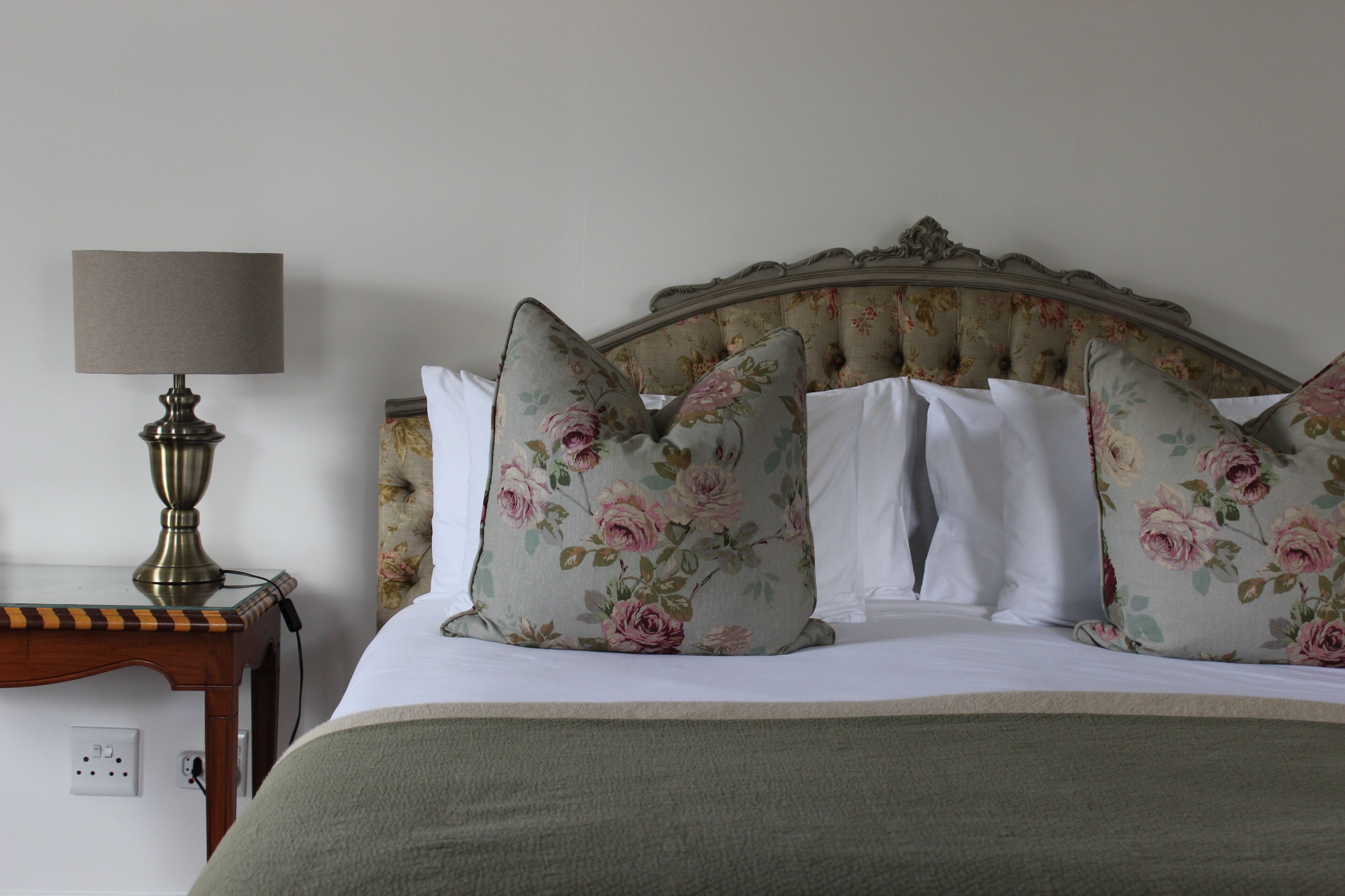 A neatly made bed with floral pillows, a bedside lamp, and a wooden nightstand with a power outlet.