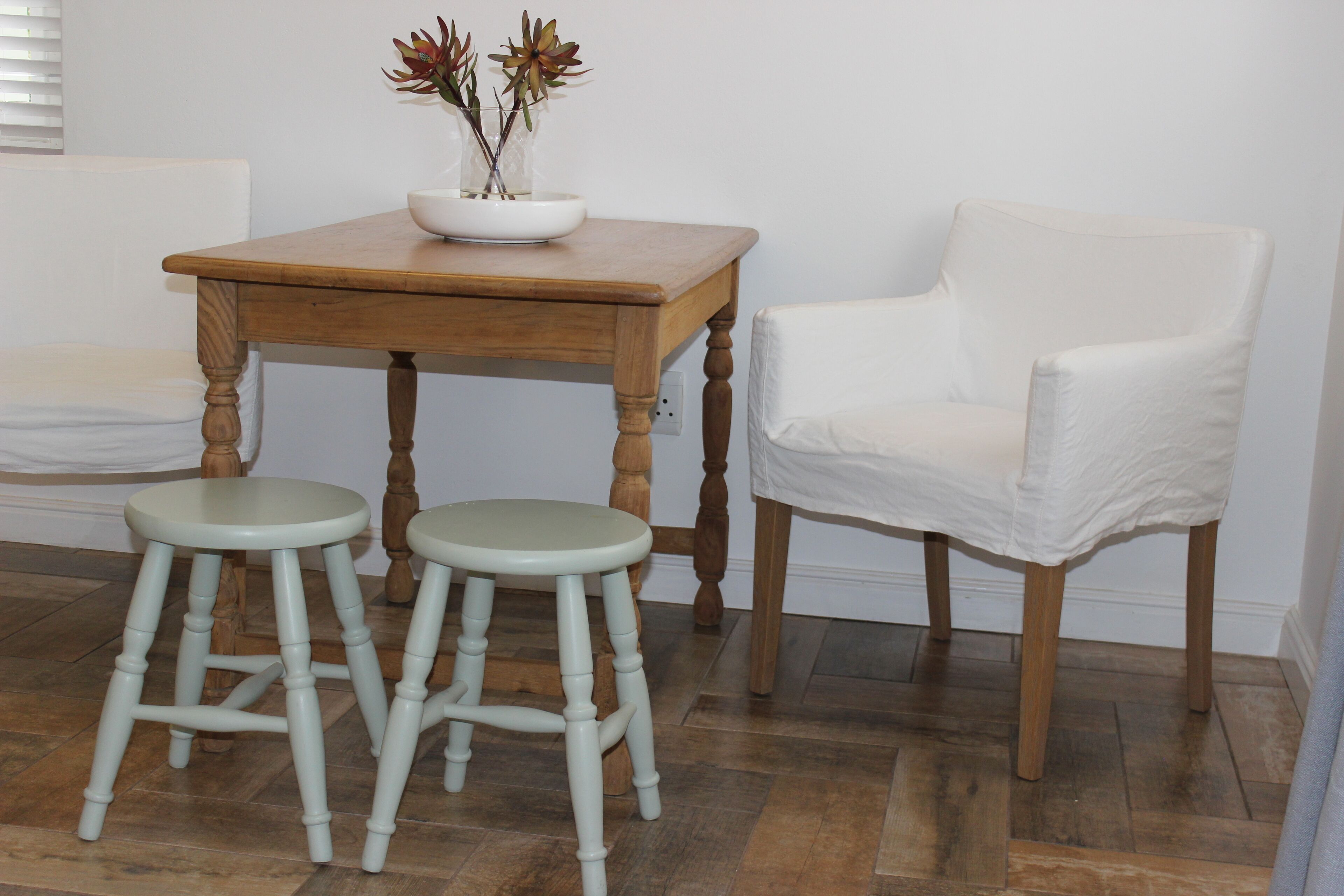 A wooden table with two stools, a white chair, and a vase with flowers.