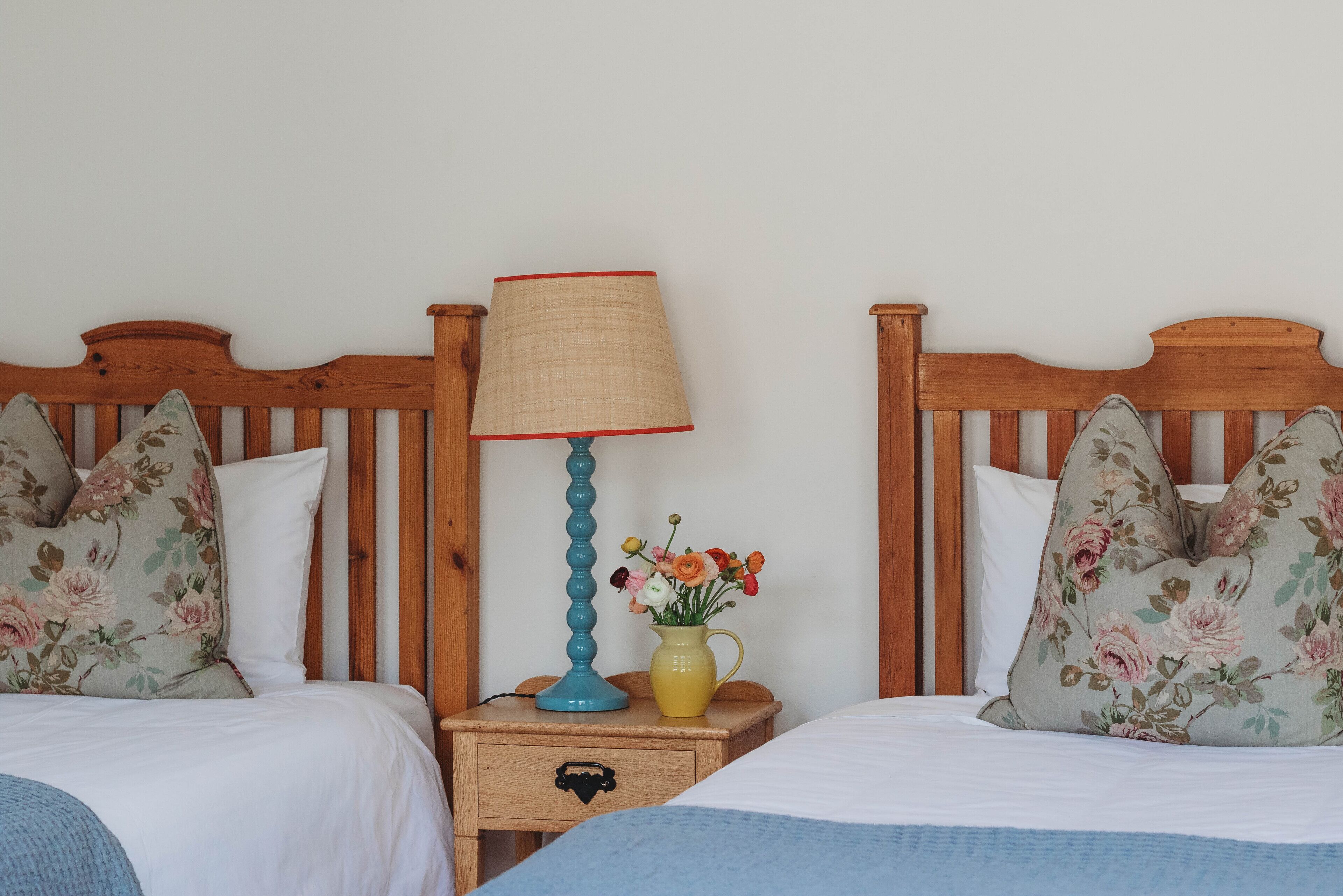 A bedroom with two wooden beds, a nightstand with a lamp and vase, and floral pillows.