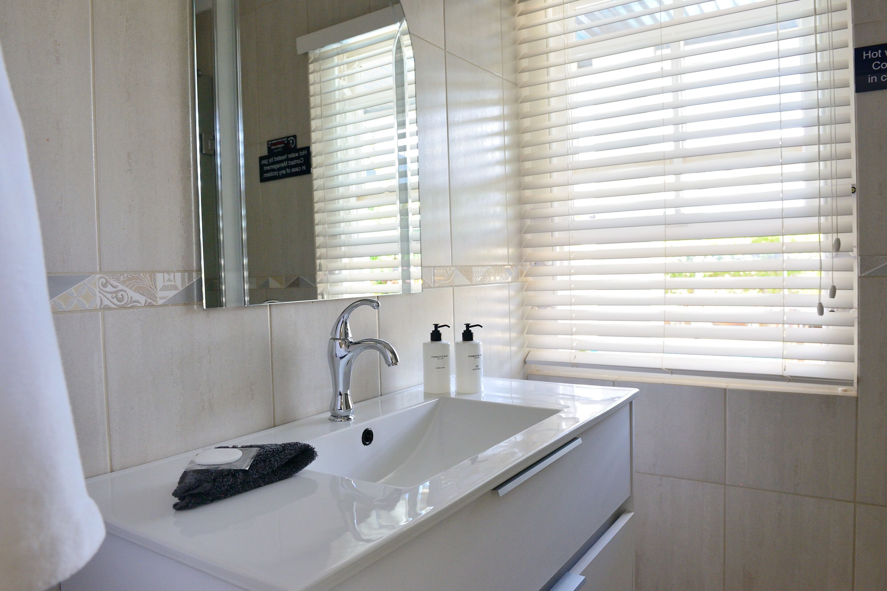 Un baño con lavamanos blanco, un espejo y una ventana con persianas.