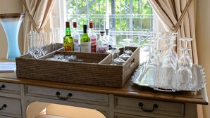 A bar area with a wicker tray holding bottles and glasses, a metal railing, and a window with curtains. - Le Petit Paris (Franschhoek)