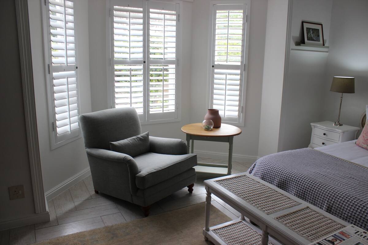 A bedroom with a bed, a chair, a small table, and a window with shutters.