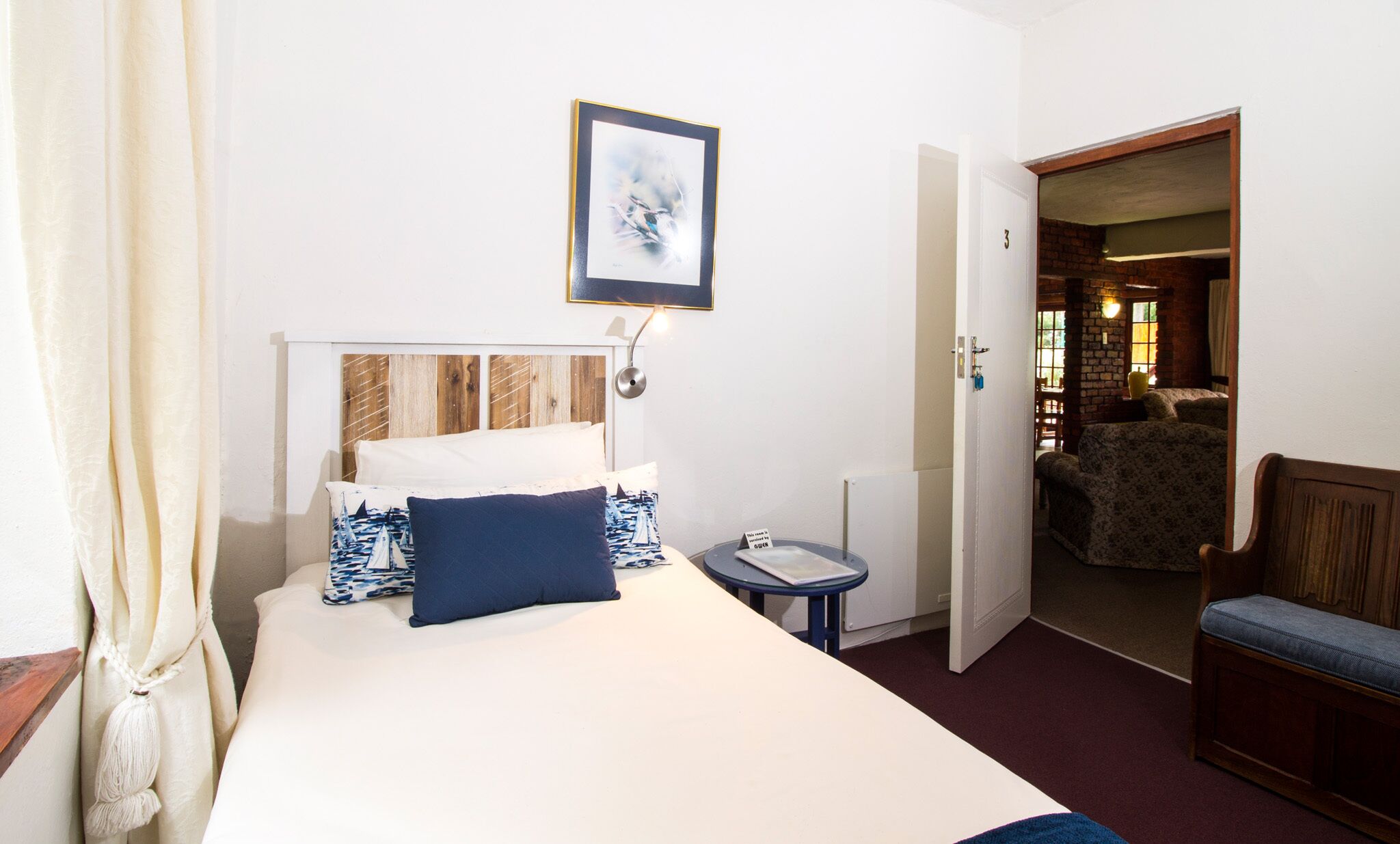 A hotel room with a bed, a bedside table, a lamp, a chair, and a door leading to another room.