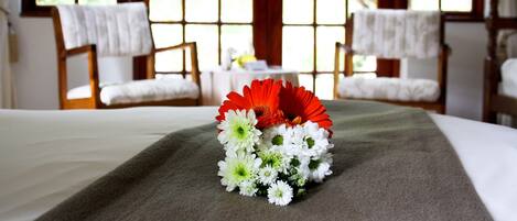 Quarto de hotel com cama, duas cadeiras, uma mesa com um vaso de flores e janelas amplas com vista para área verde.