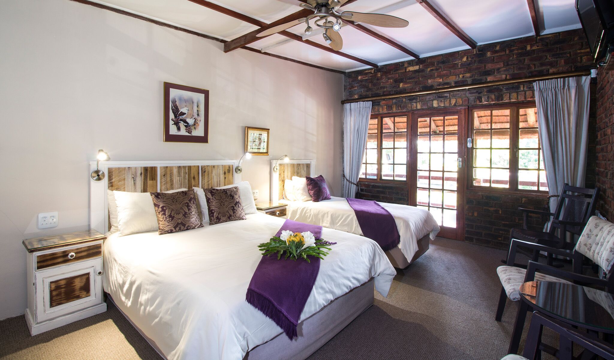 A hotel room with two beds, a ceiling fan, a large window with curtains, and a chair.
