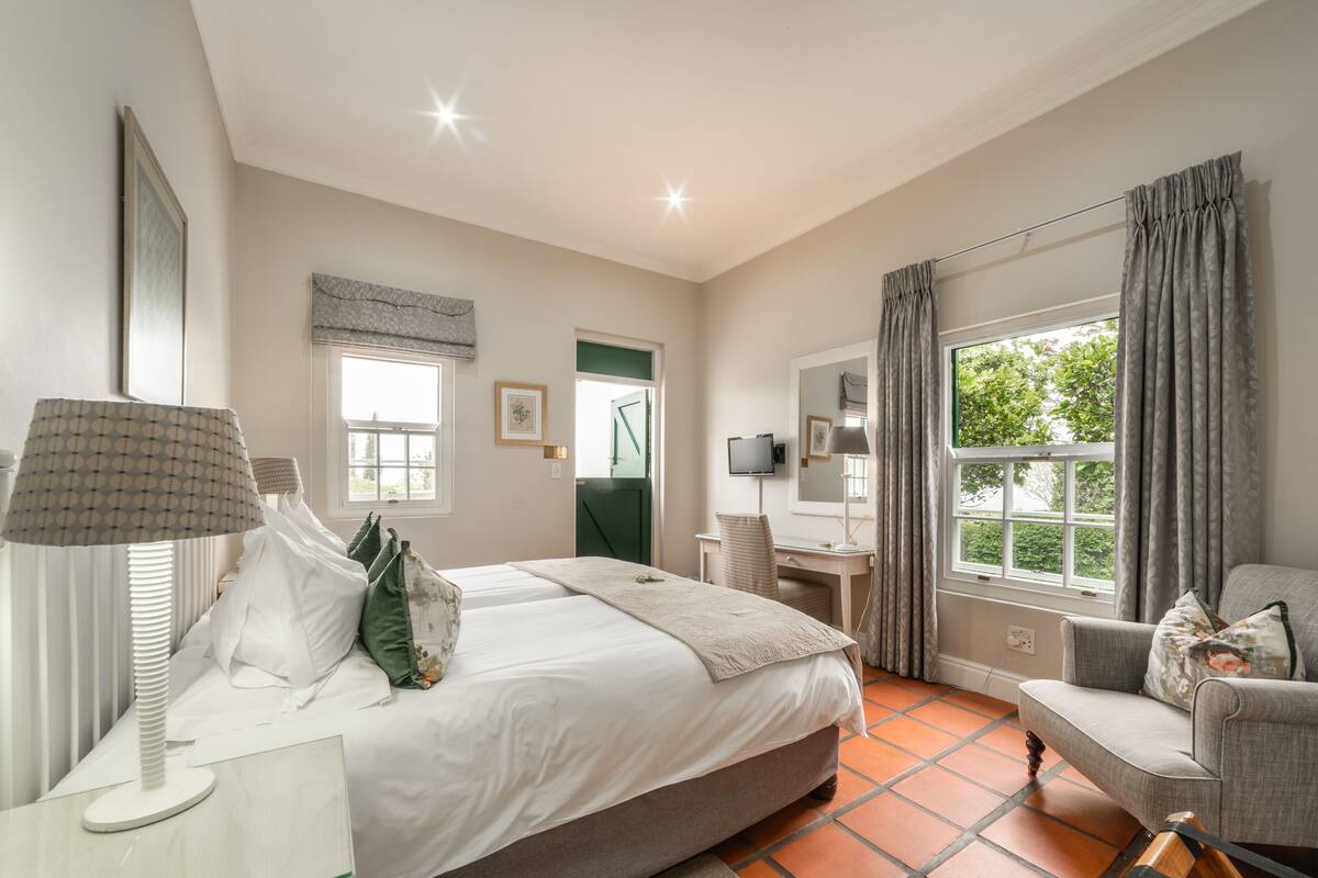 Un dormitorio con cama, mesita de noche, lámpara, silla, escritorio, televisor y ventana con cortinas.