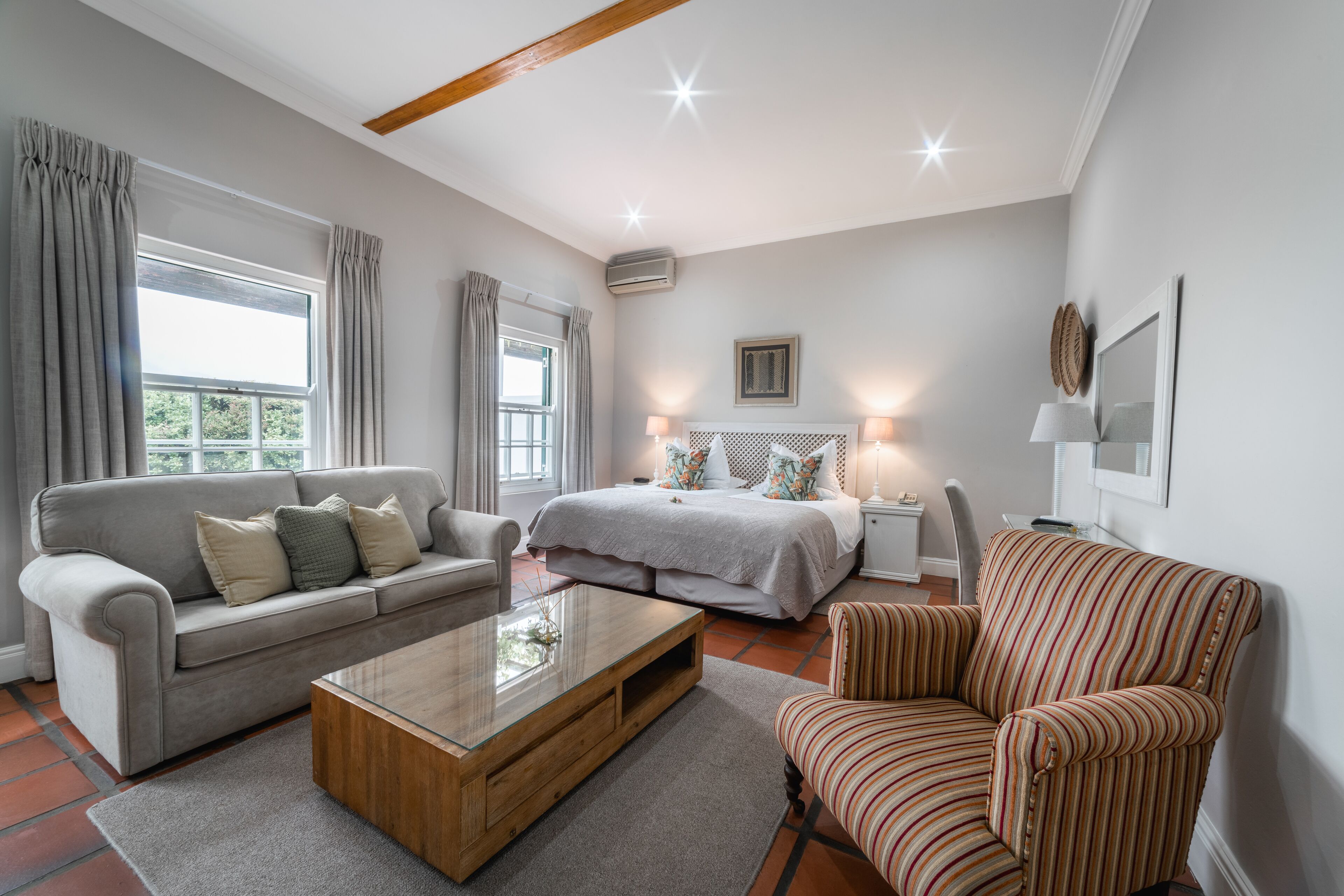 A spacious living room with a sofa, a coffee table, a striped armchair, a bed with a headboard, and a small side table with a lamp.
