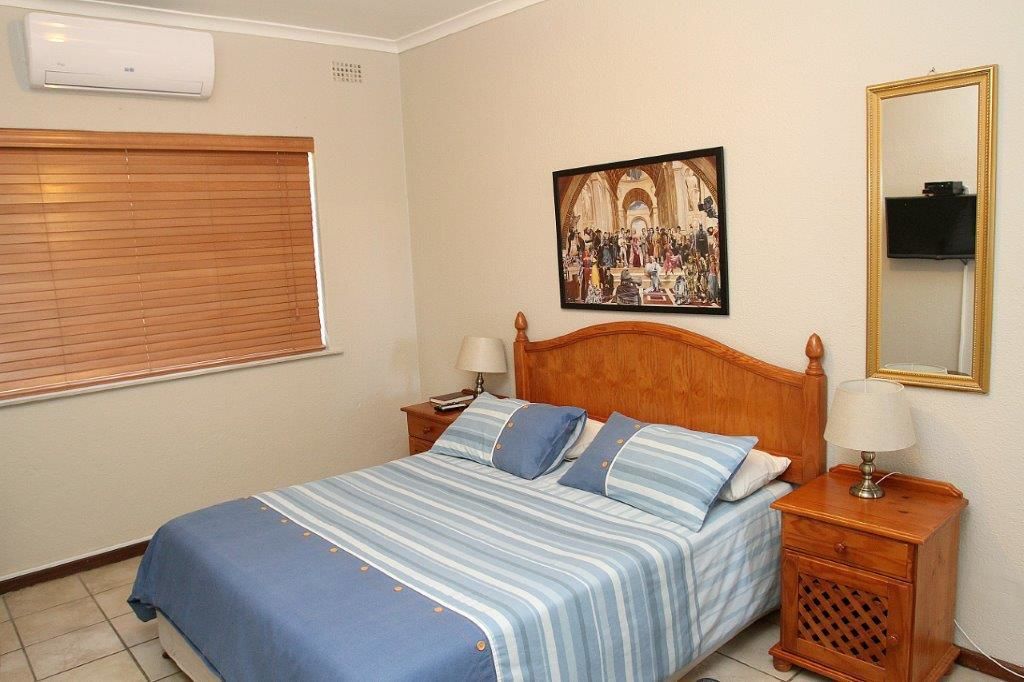A bedroom with a bed, two nightstands, a wall-mounted air conditioner, a painting, and a mirror.