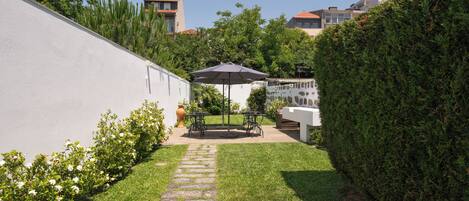 A backyard with a stone pathway leading to a table and chairs under a large umbrella, surrounded by well-maintained grass and plants.