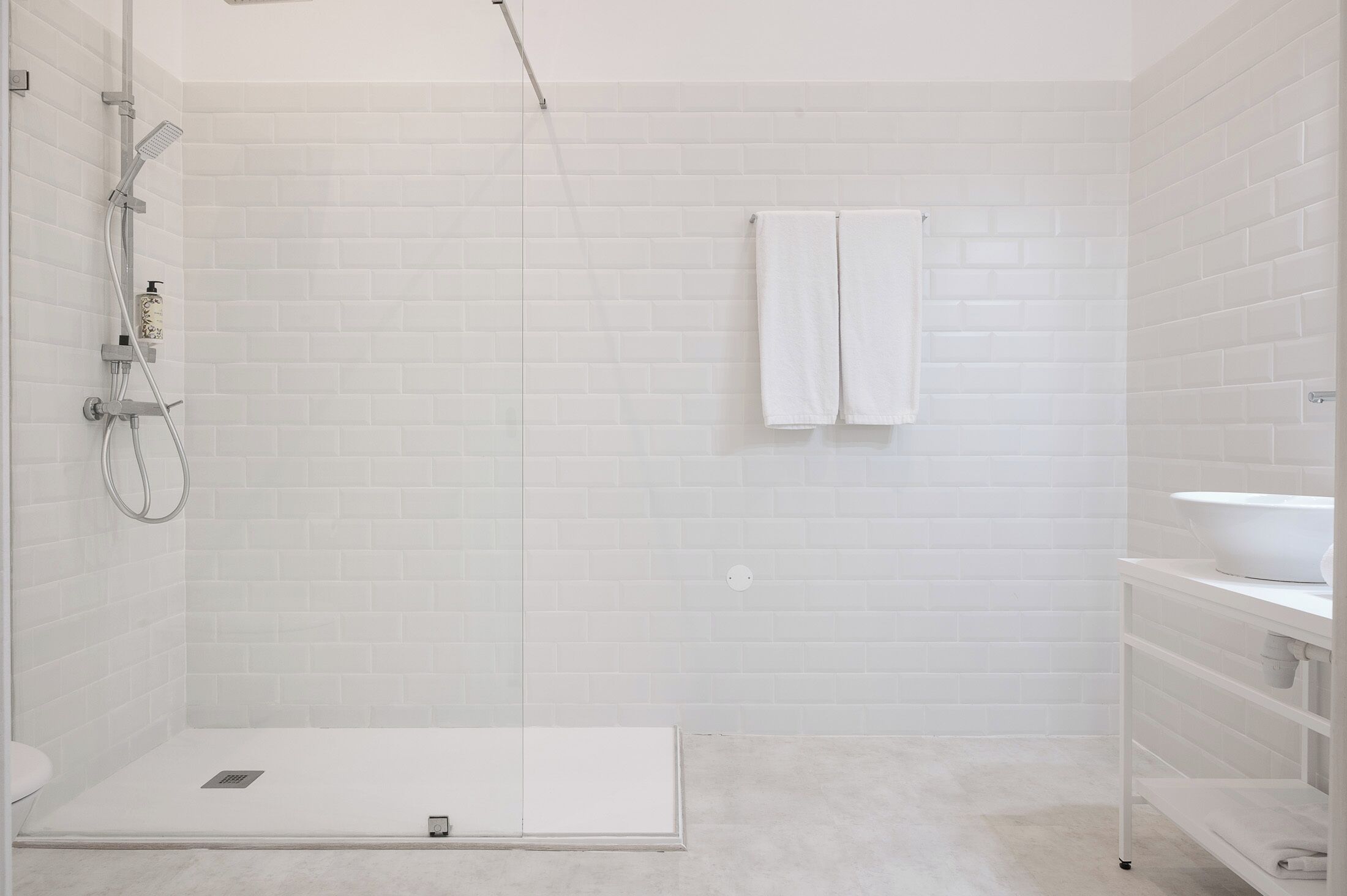 Un baño moderno con azulejos blancos tipo metro, ducha con mampara de vidrio, lavamanos blanco y un perchero.