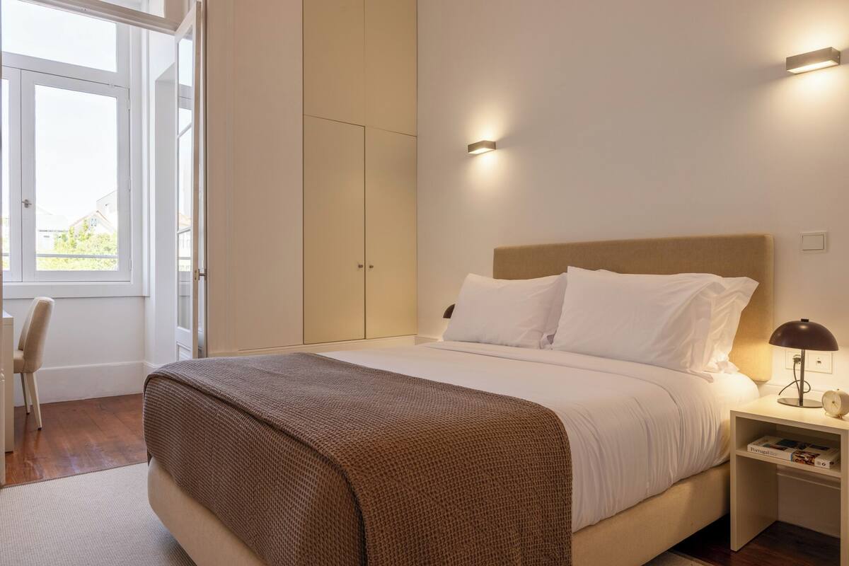A neatly made bed with white linens and a brown throw blanket, a bedside table with a lamp, and a window with a view of greenery outside.