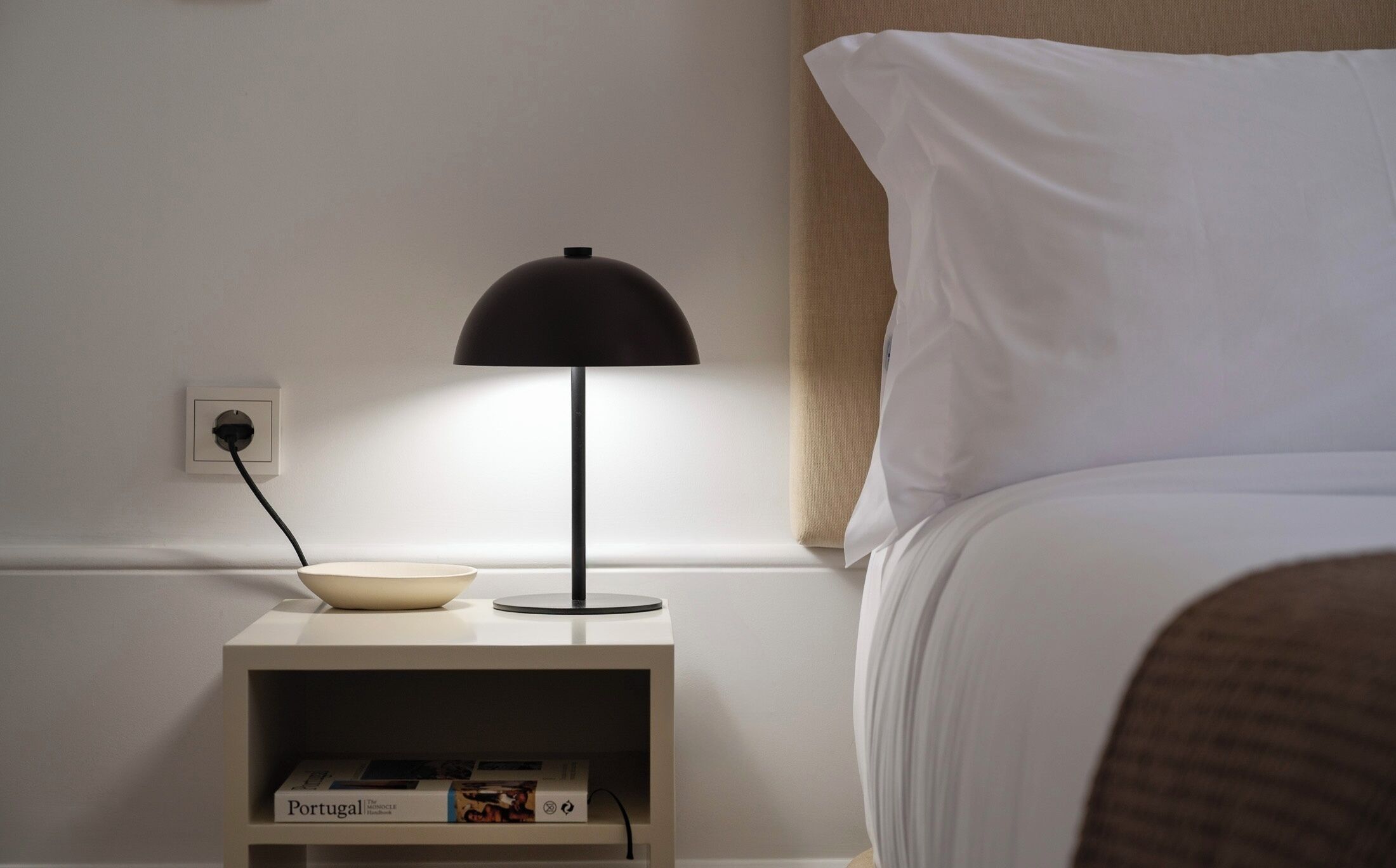 A neatly made bed with white bedding, a bedside table with a black lamp, and a book titled "Portugal" on the shelf.