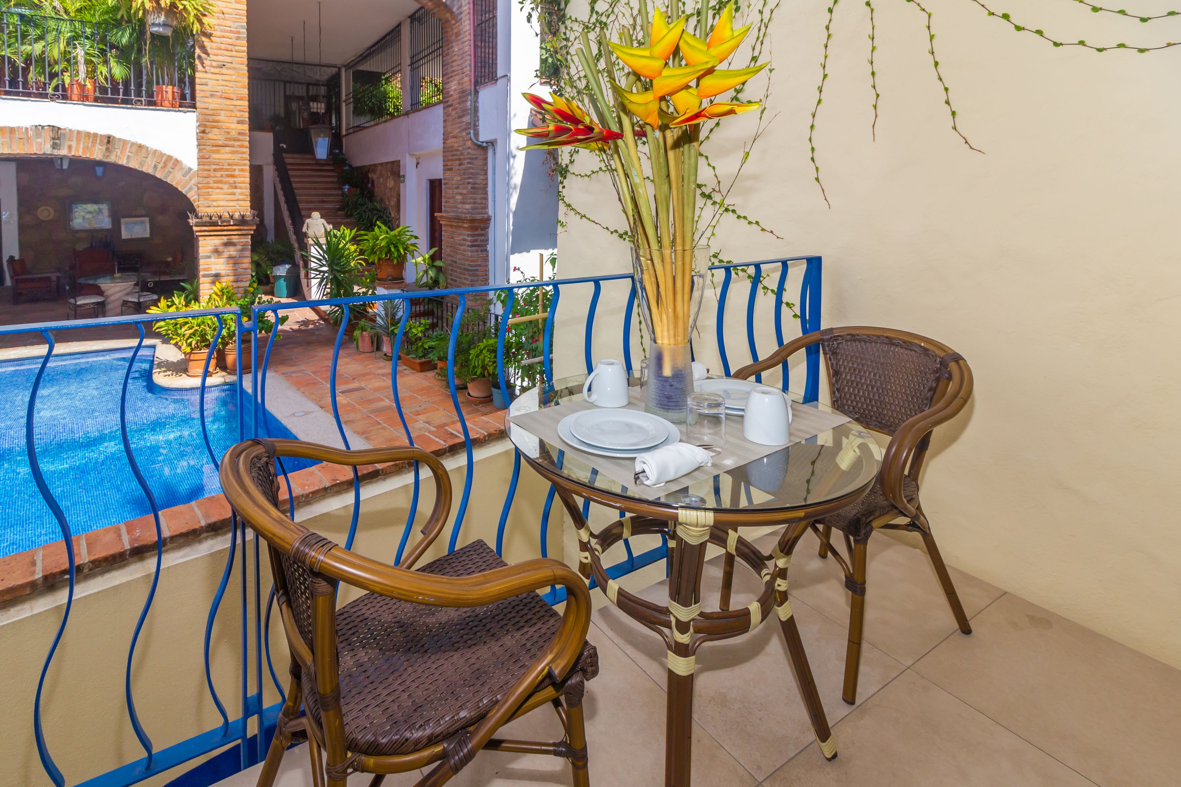 A balcony with a glass table, two wicker chairs, a vase with flowers, and a pool in the background.