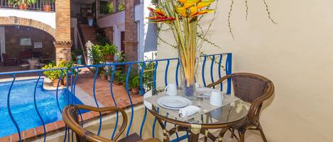 A balcony with a glass table, two wicker chairs, a vase with flowers, and a pool in the background.