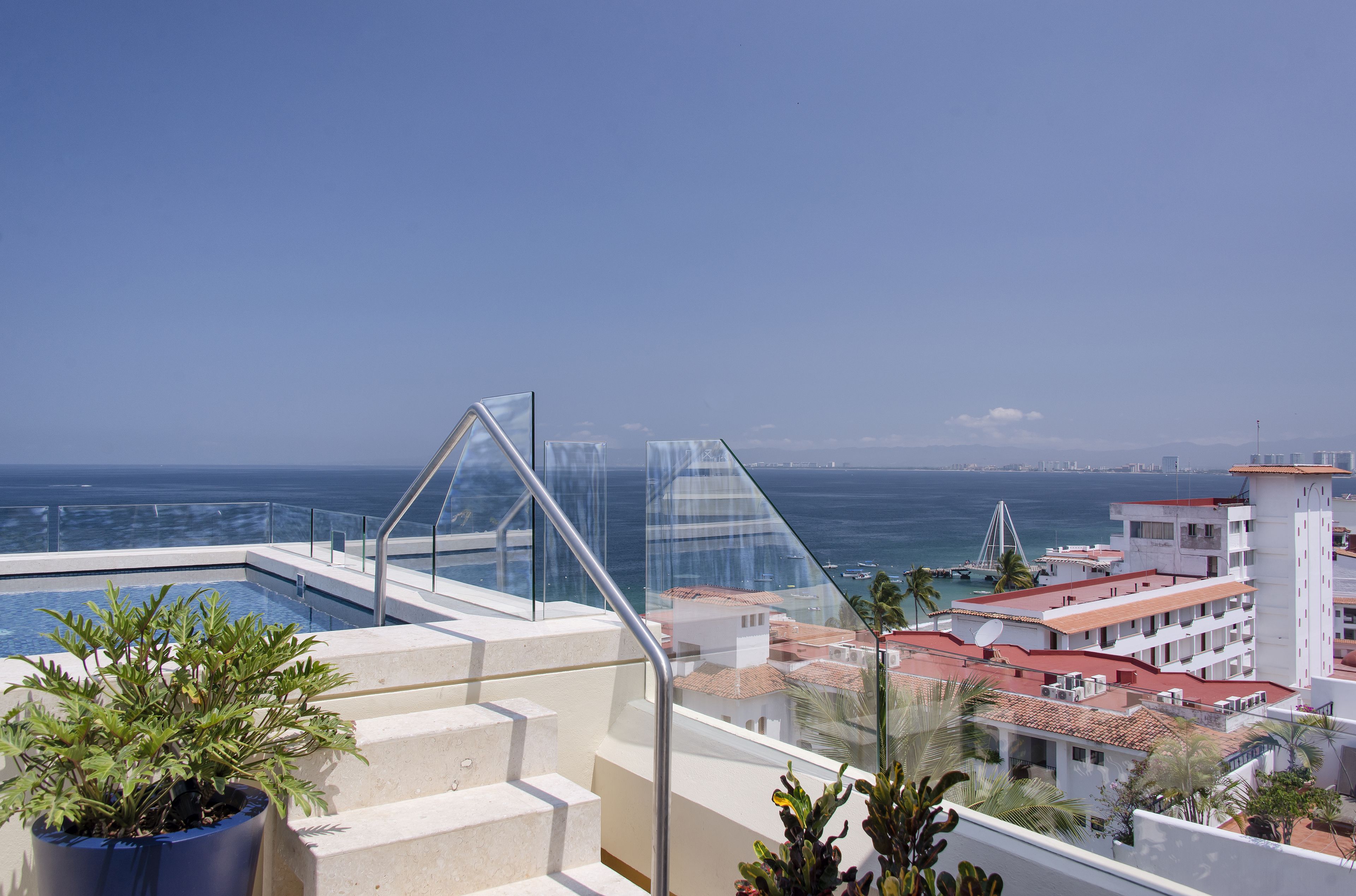 A rooftop pool with a glass balustrade, a clear view of the ocean, and surrounding buildings with red-tiled roofs.