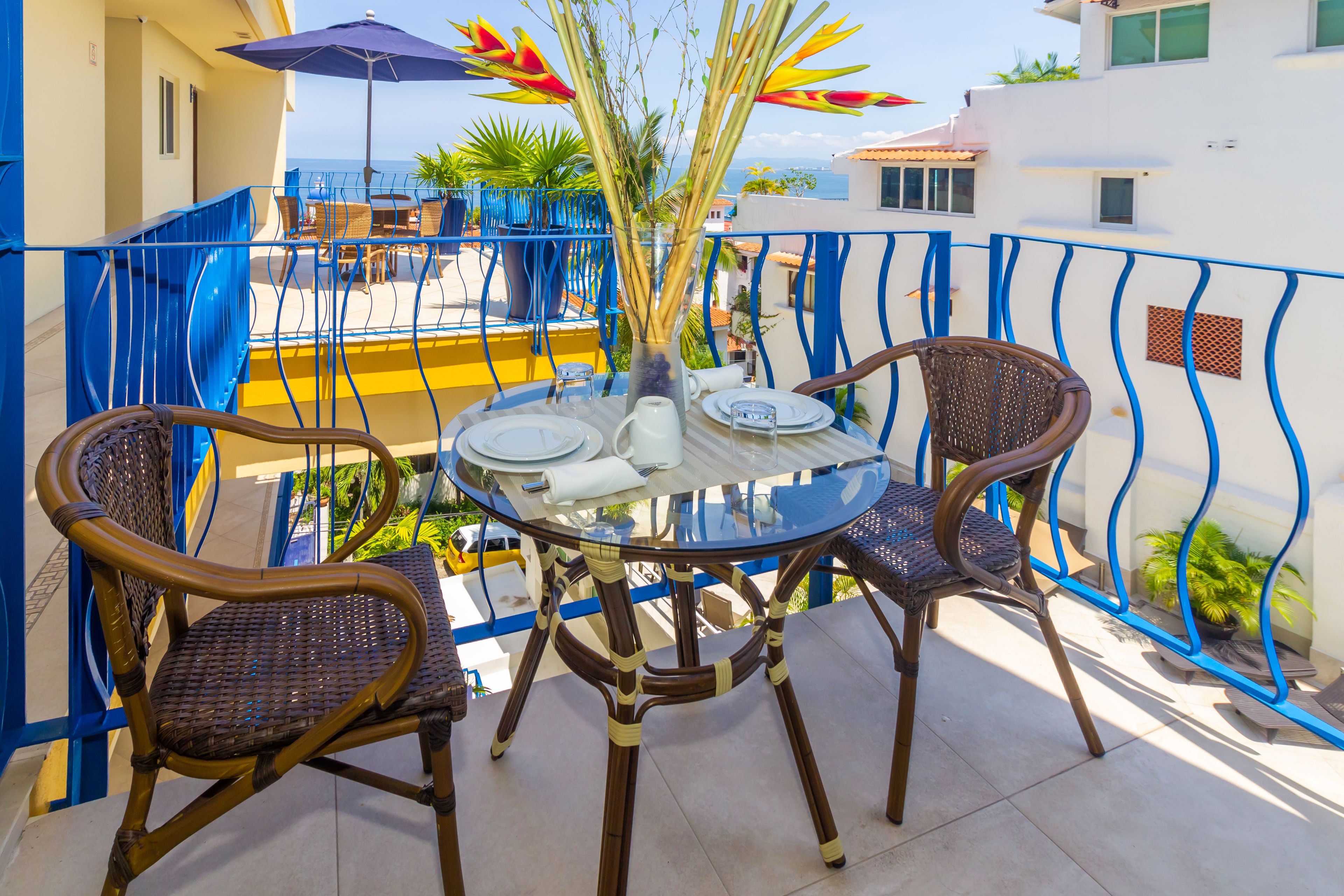Una mesa redonda de cristal con cuatro sillas de mimbre, un jarrón con plantas y un paraguas azul en un balcón con vistas al mar.