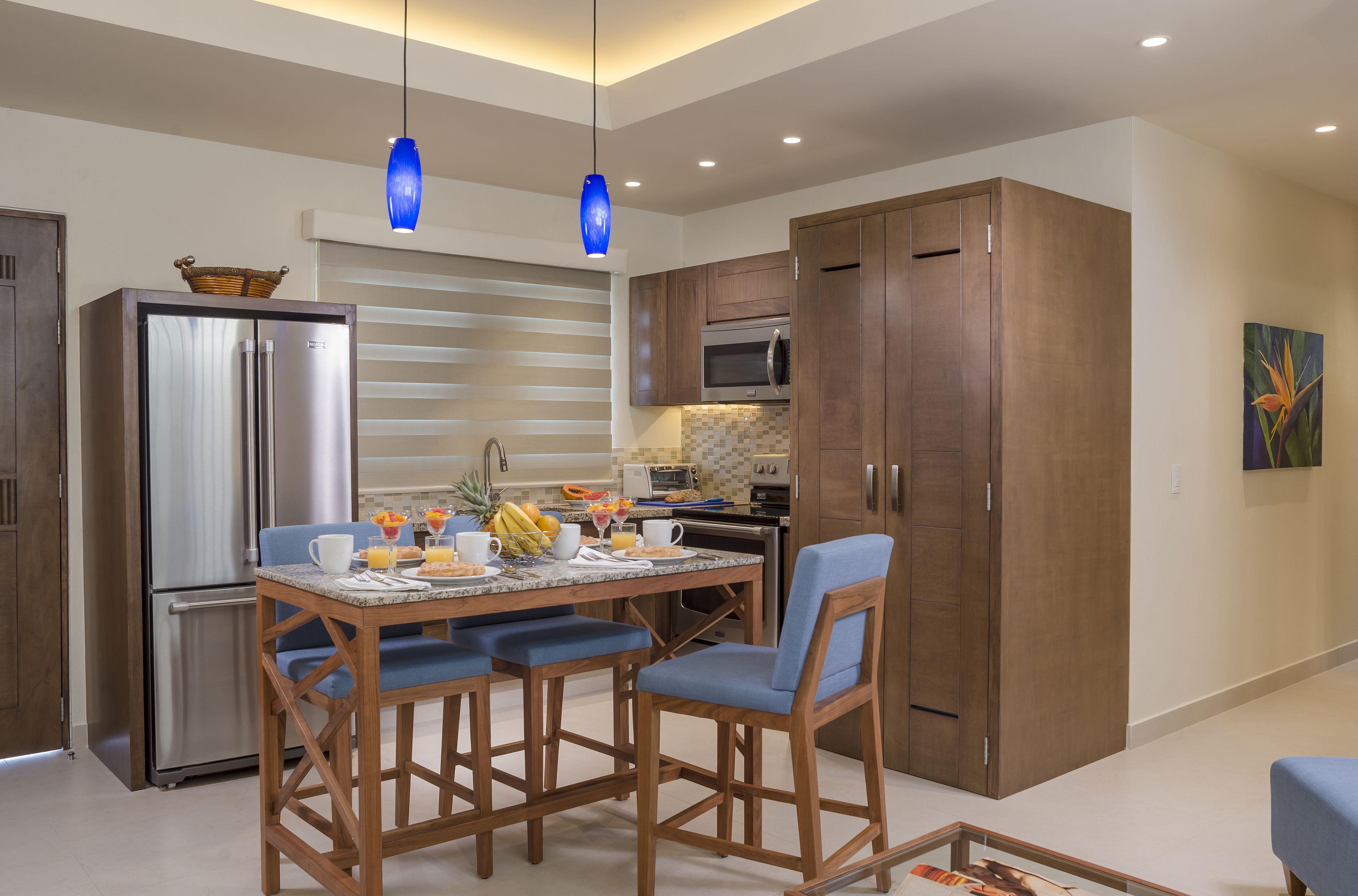Une cuisine moderne dotée d’un réfrigérateur en acier inoxydable, de meubles en bois et d’un coin repas avec des chaises bleues et un comptoir en marbre.