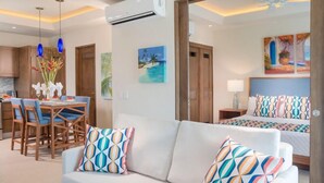 Una habitación de hotel moderna con sofá, una mesa de comedor con sillas, cama con ropa de cama colorida y obras de arte en las paredes.