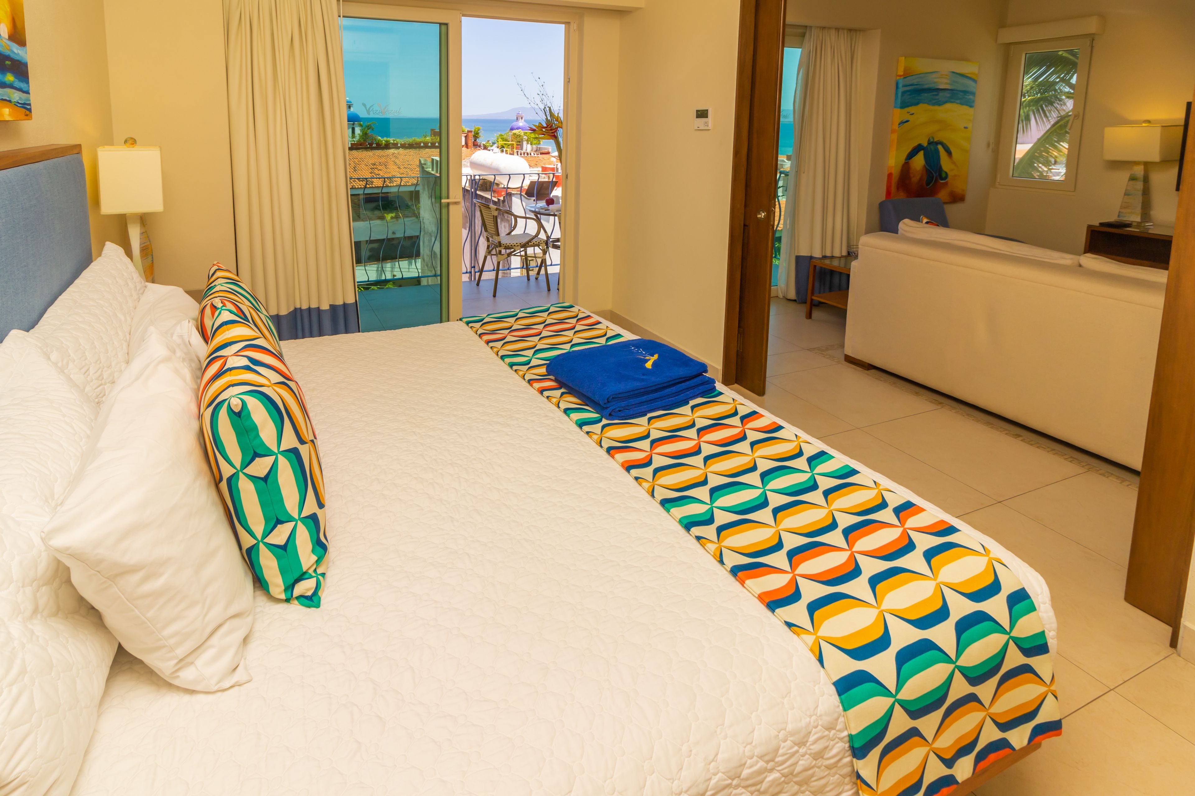 A hotel room with a bed, a sofa, a dining area with a table and chairs, and a view of the ocean.