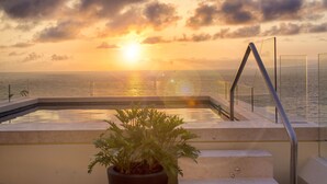 Una piscina con vista al atardecer sobre el océano, una planta en maceta y una escalera que lleva a un balcón.