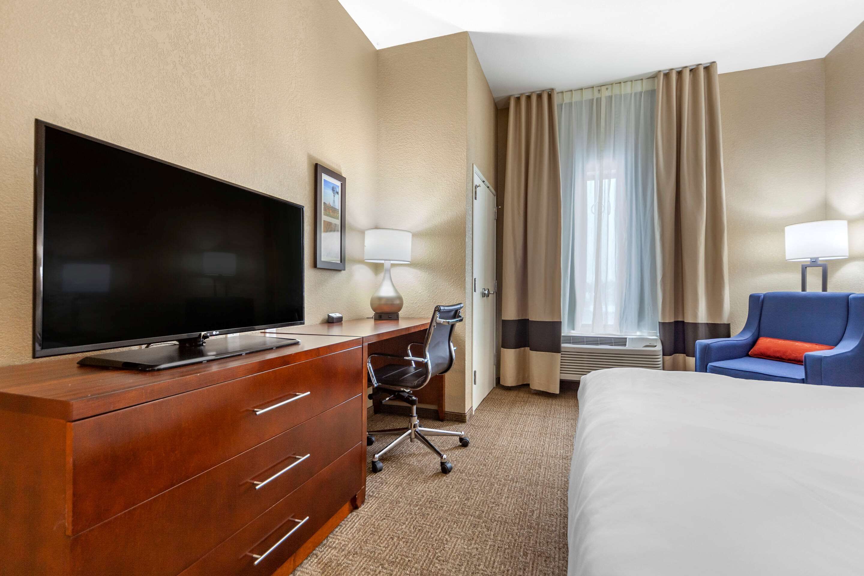A hotel room with a flat-screen TV, a wooden dresser, a desk with a chair, a bed, and a blue armchair.
