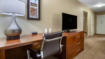 A hotel room with a wooden desk, a black office chair, a flat-screen TV, a lamp, and a framed picture on the wall.
