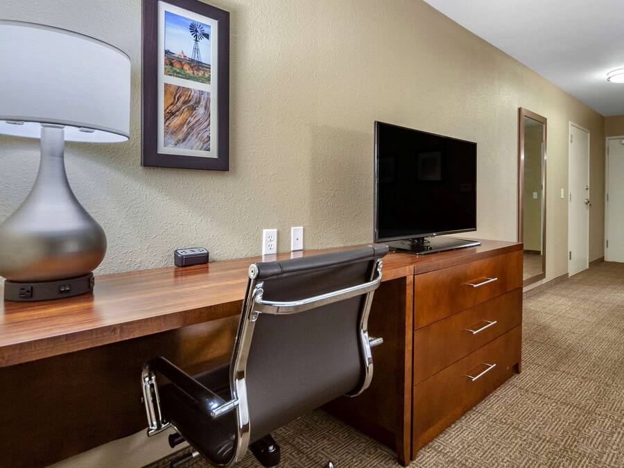 A hotel room with a wooden desk, a black office chair, a flat-screen TV, a lamp, and a framed picture on the wall.