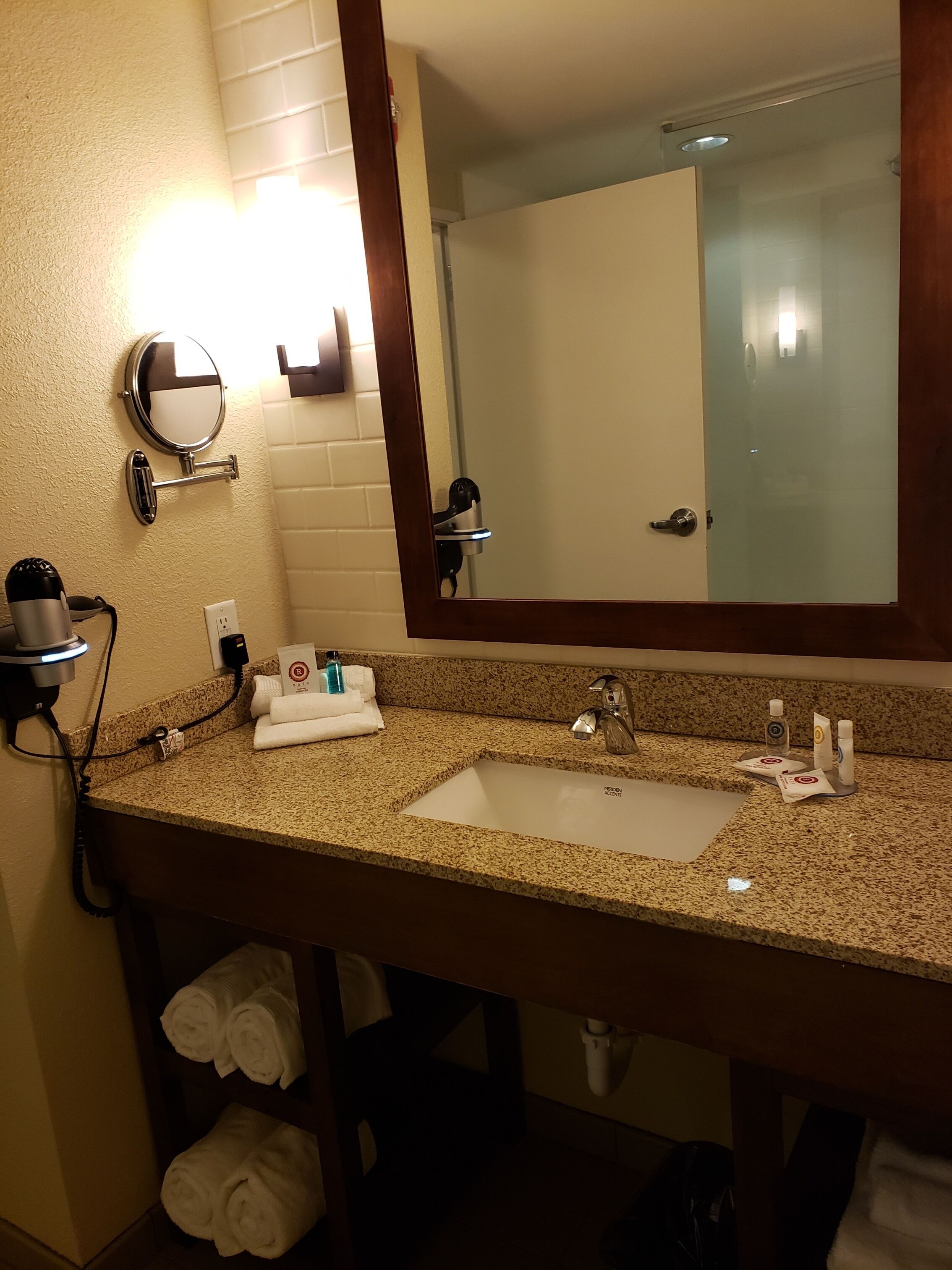 A hotel bathroom with a granite countertop sink, a mirror, a towel rack, and a hairdryer.