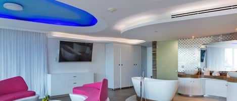 A modern hotel room with a curved ceiling, a large flat-screen TV, a white bathtub, and a pink and white sofa set.