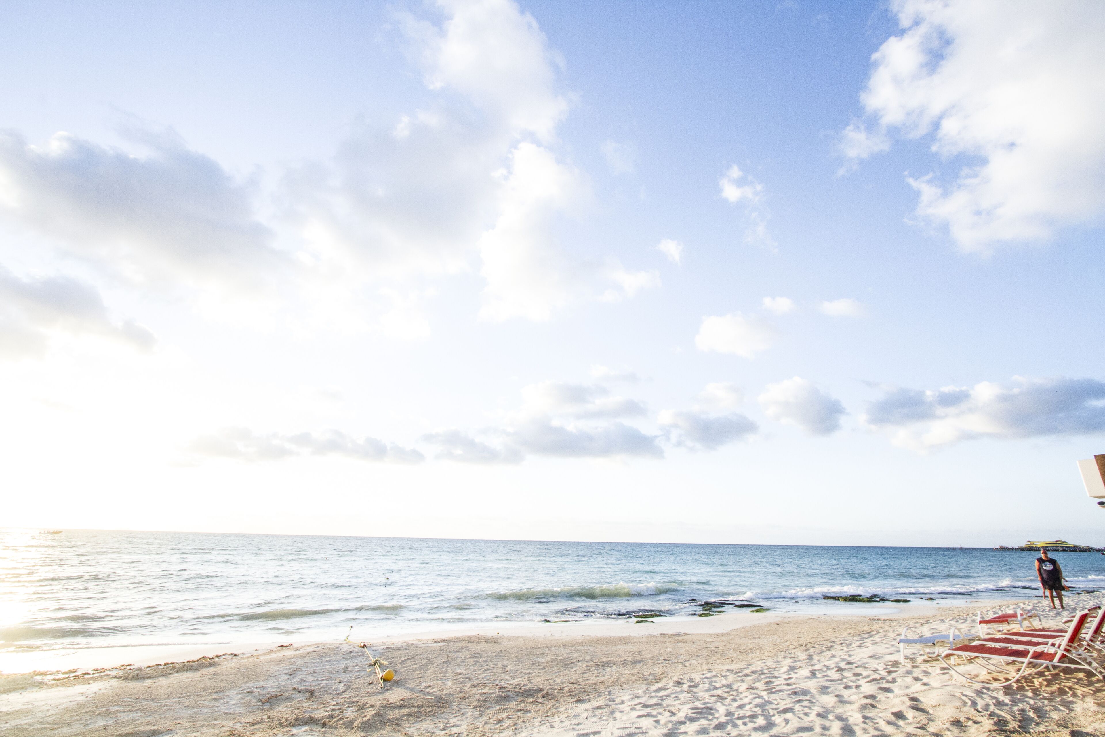 A beach with a person walking, red lounge chairs, and a clear sky.