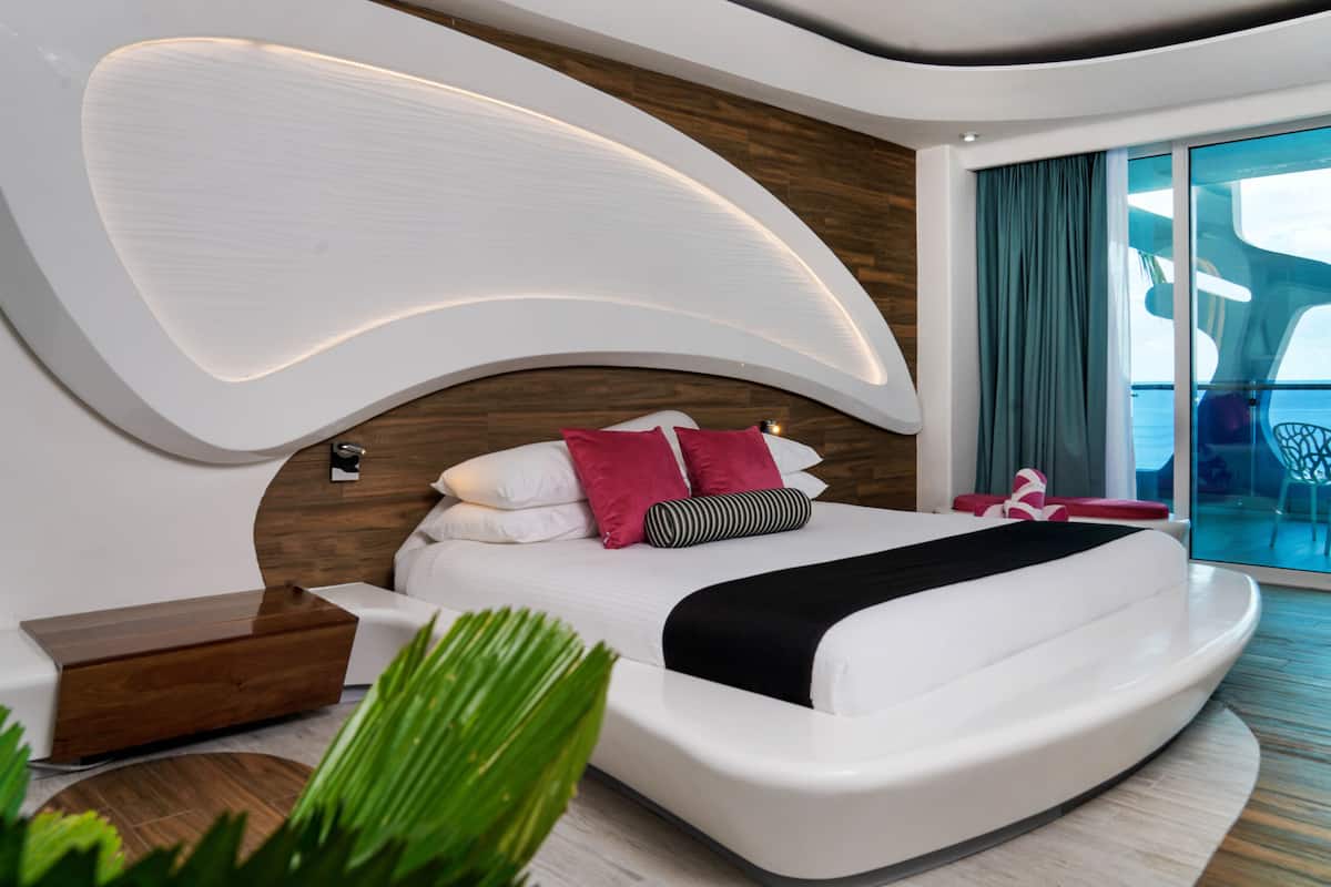 Ein modernes Schlafzimmer mit einer gebogenen Kopfteil, einem großen Bett mit weißer Bettwäsche und roten Kissen, einem Nachttisch aus Holz und Blick auf das Meer durch große Fenster.