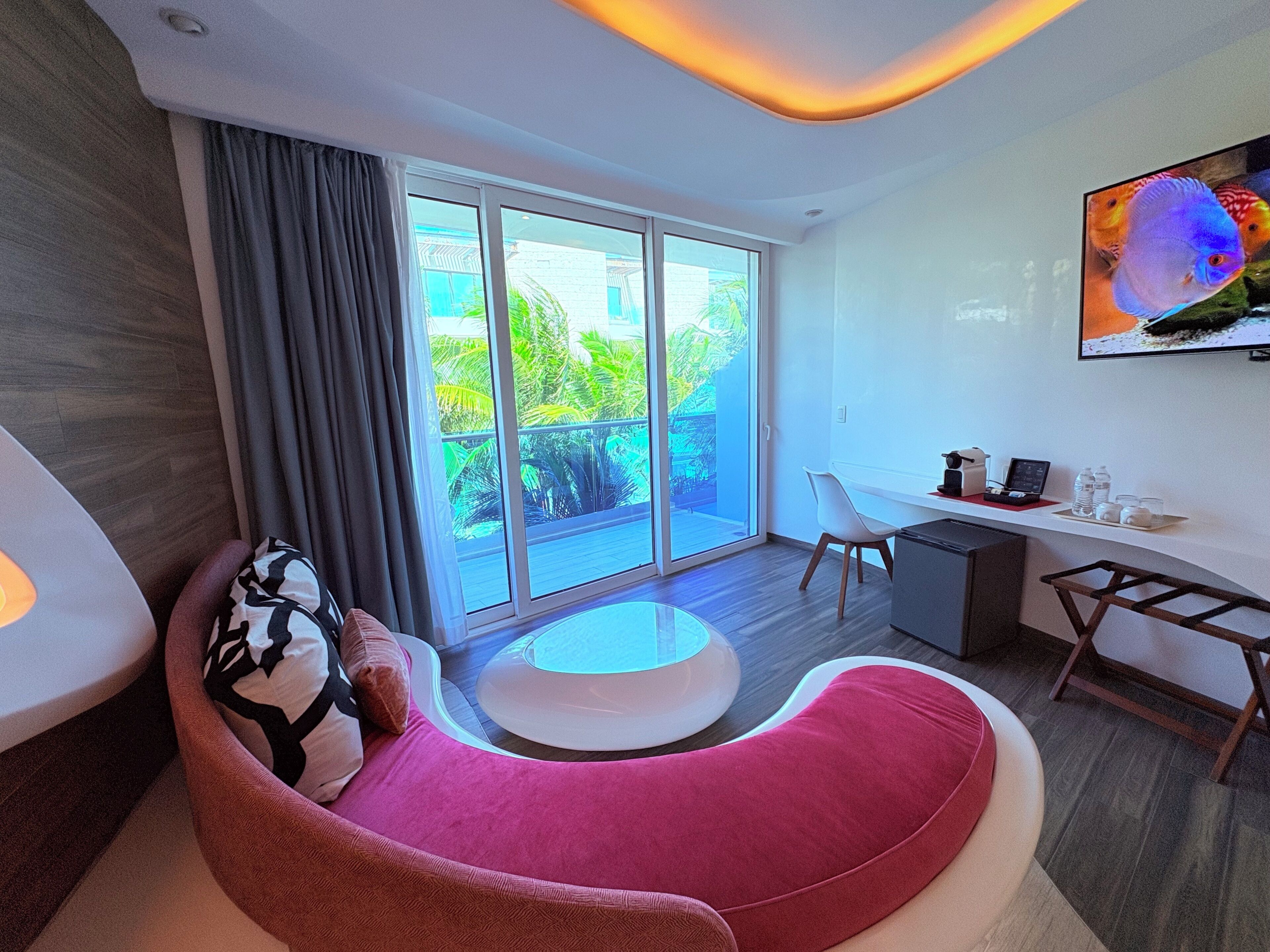 A modern hotel room with a curved sofa, a desk, and a large window offering a view of greenery.