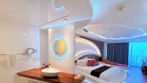 Une chambre d’hôtel moderne dotée d’un lavabo central, d’un miroir rond, d’un lit avec une literie blanche et d’un plafond aux lignes courbes.