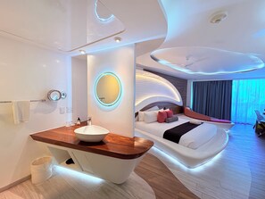Una habitación de hotel moderna con un lavamanos central tipo isla, un espejo redondo, una cama con ropa blanca y un diseño de techo curvo.