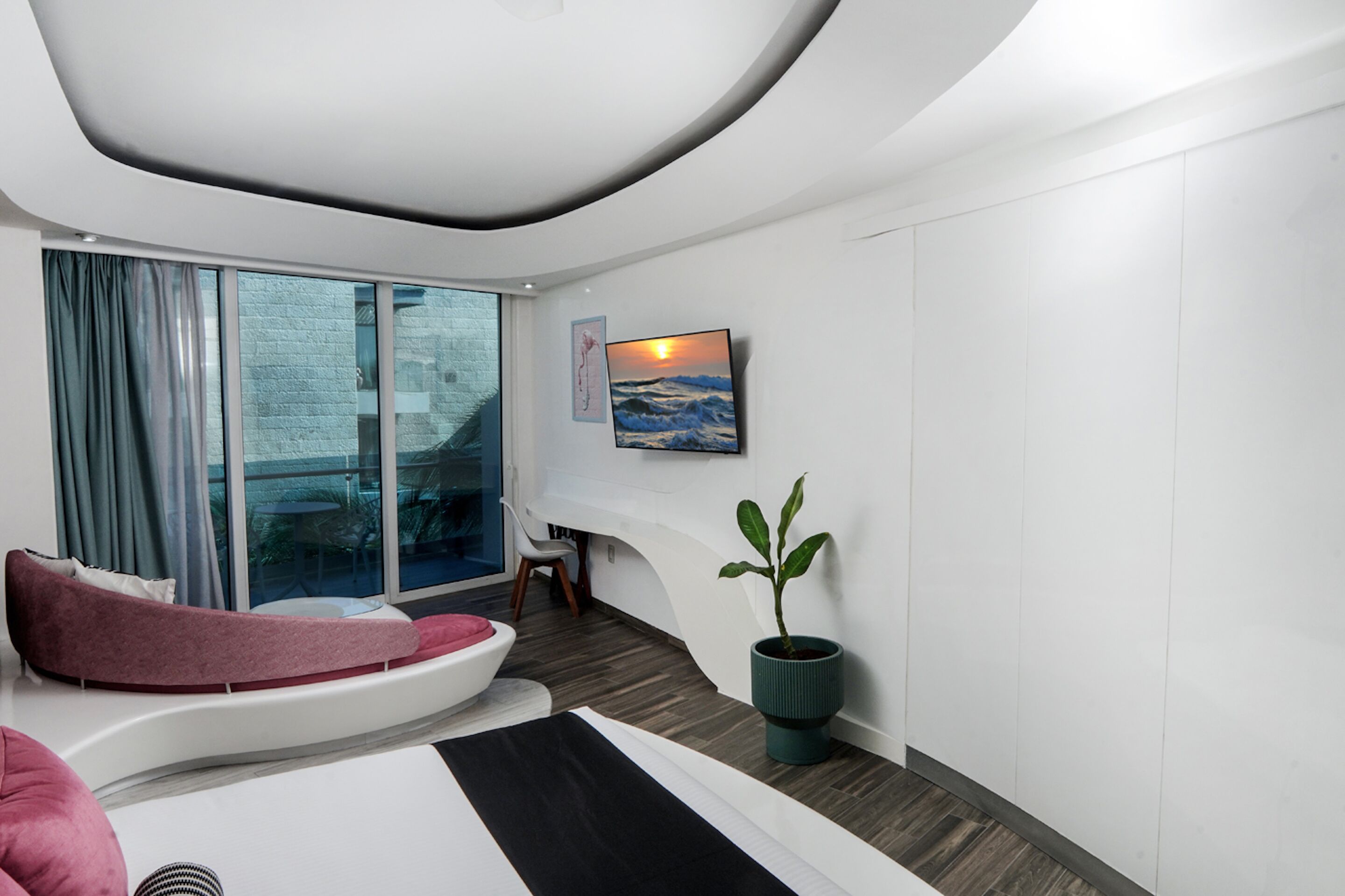 Une chambre d’hôtel moderne avec un plafond incurvé, une grande fenêtre avec des rideaux, un lit doté d’une tête de lit rose, une plante en pot et un tableau accroché au mur.