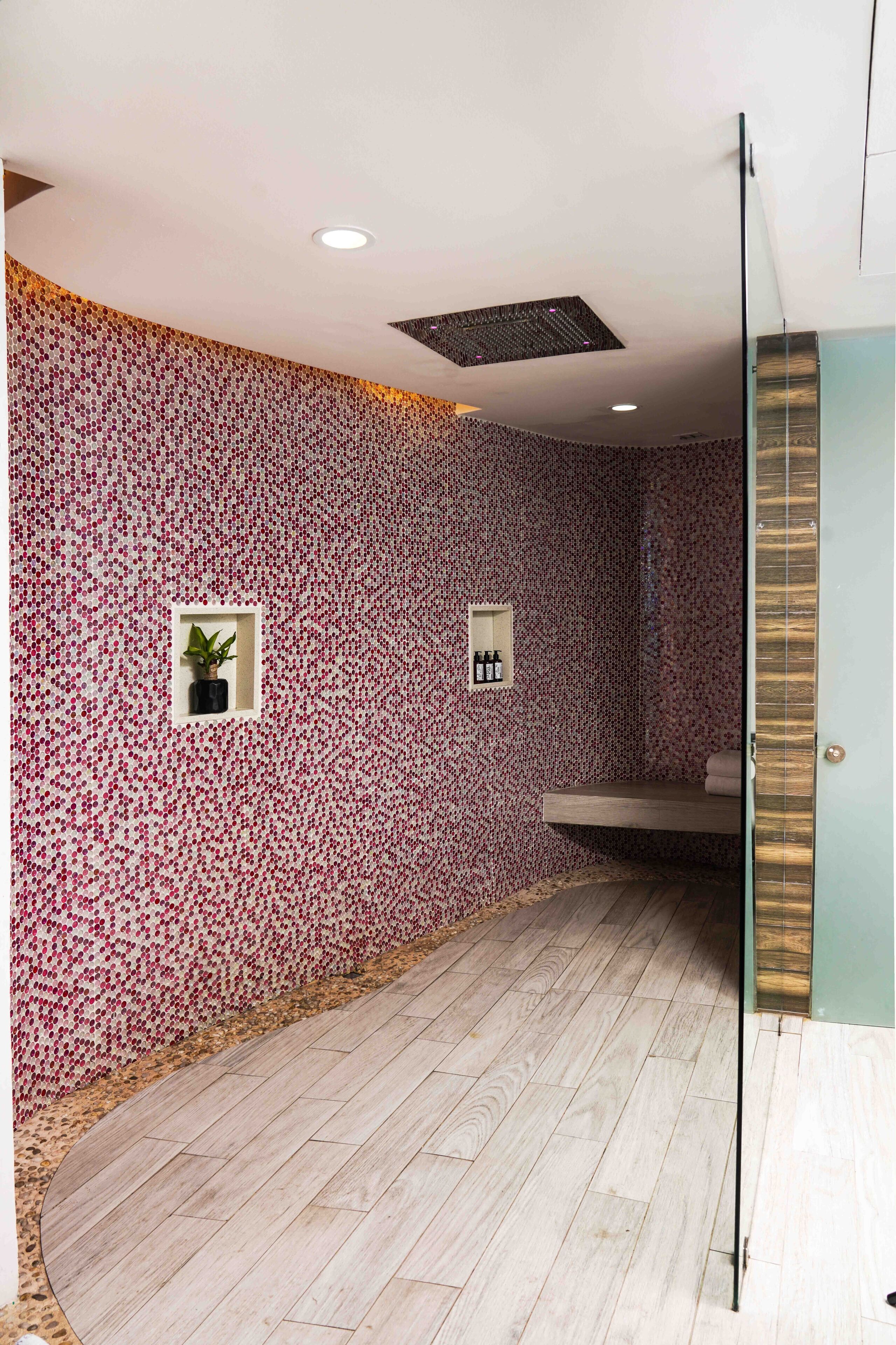 A bathroom with a patterned wall, a wooden floor, a shelf with toiletries, and a small plant.