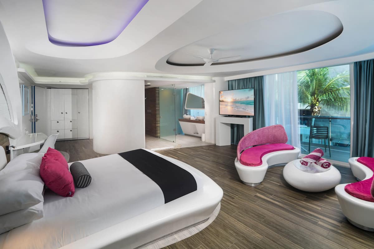 Ein modernes Hotelzimmer mit einem großen Bett, zwei rosafarbenen Sessel, einem weißen Sofa, einem Fernseher und einem Balkon mit Blick auf Palmen.