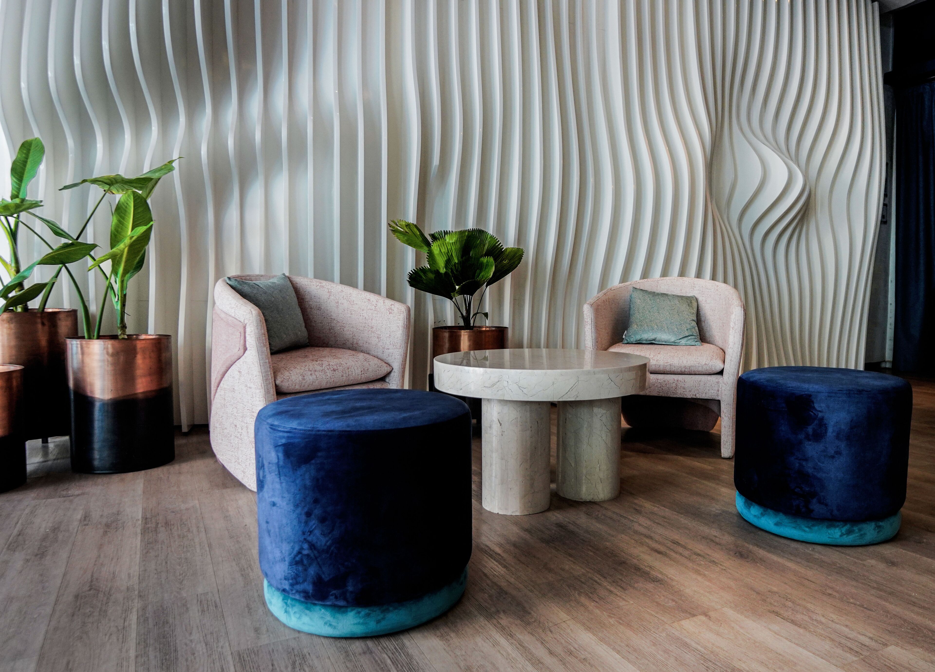 A modern lounge area with two plush armchairs, a round coffee table, and two round ottomans. There are potted plants and a textured wall in the background.