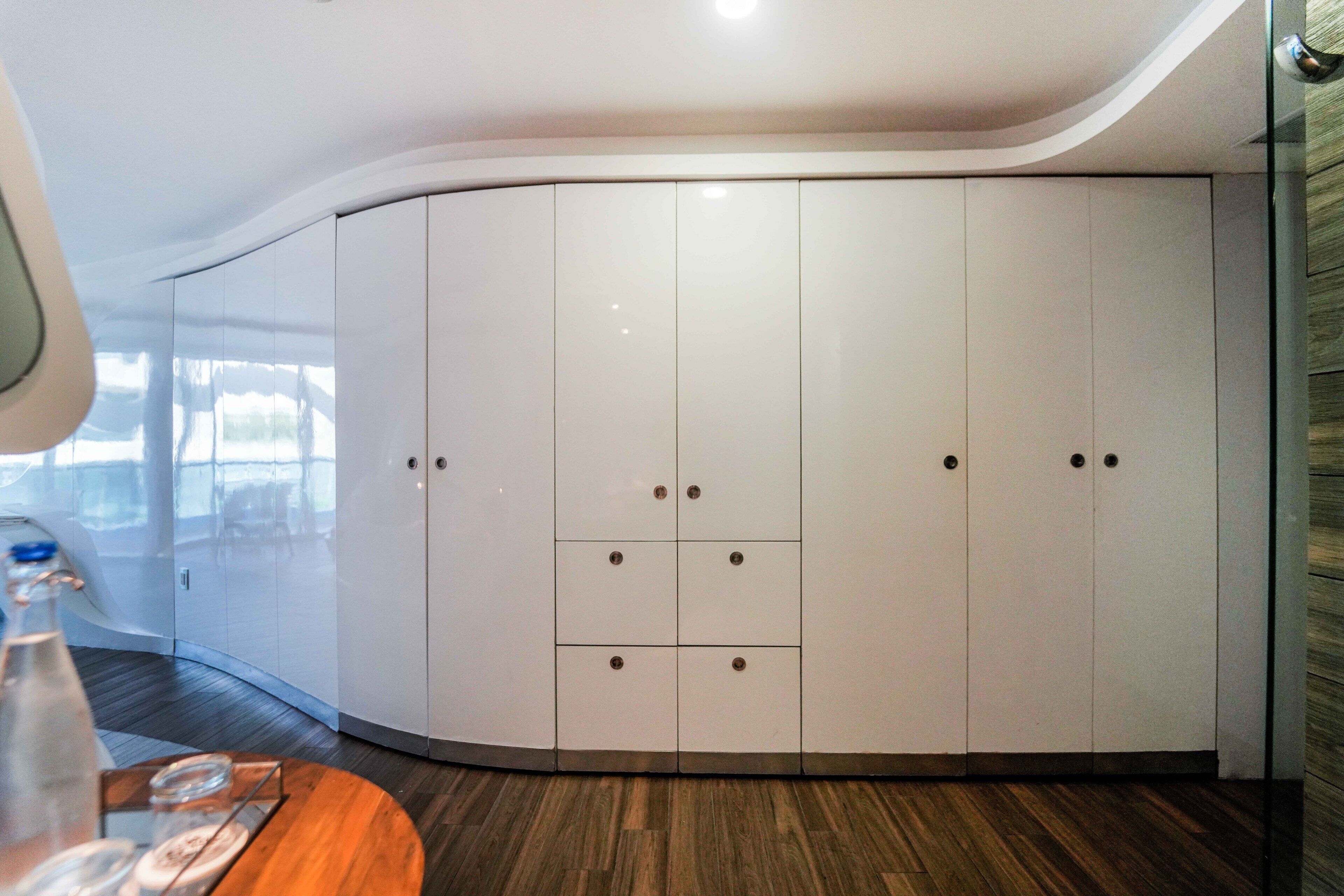 A modern room with a curved white wardrobe, wooden flooring, and a glass table with a bottle and glasses.