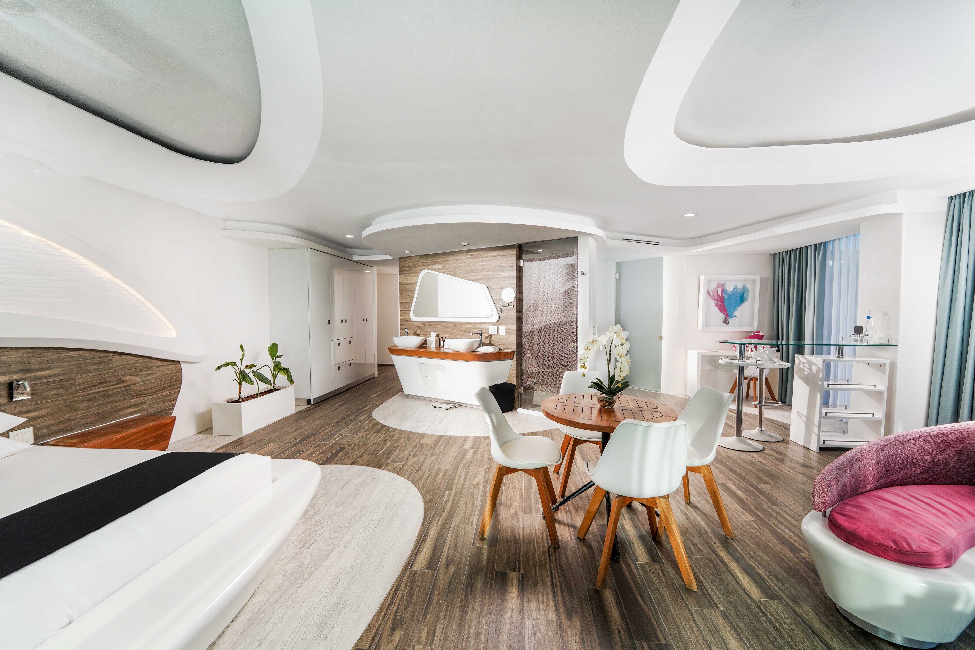 A modern hotel room with a large bed, a dining area with a round table and chairs, a small kitchen, and a living area with a pink sofa.