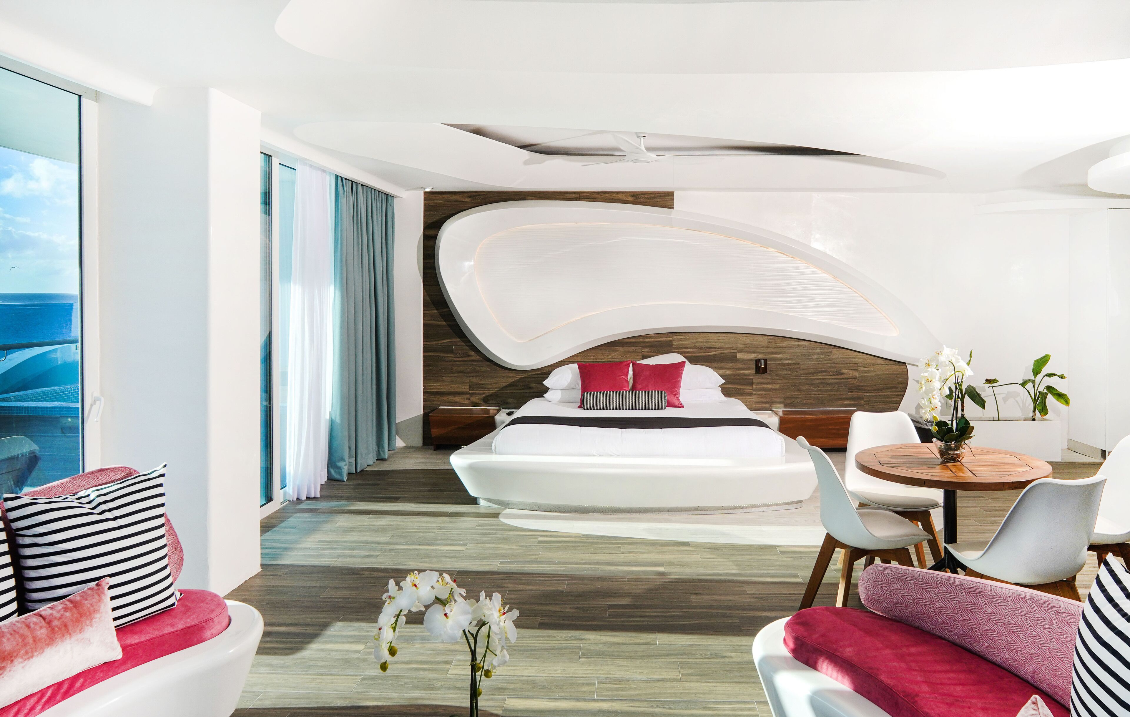 A modern hotel room with a curved ceiling, a large bed with red and black pillows, a round dining table with chairs, and a view of the ocean through large windows.