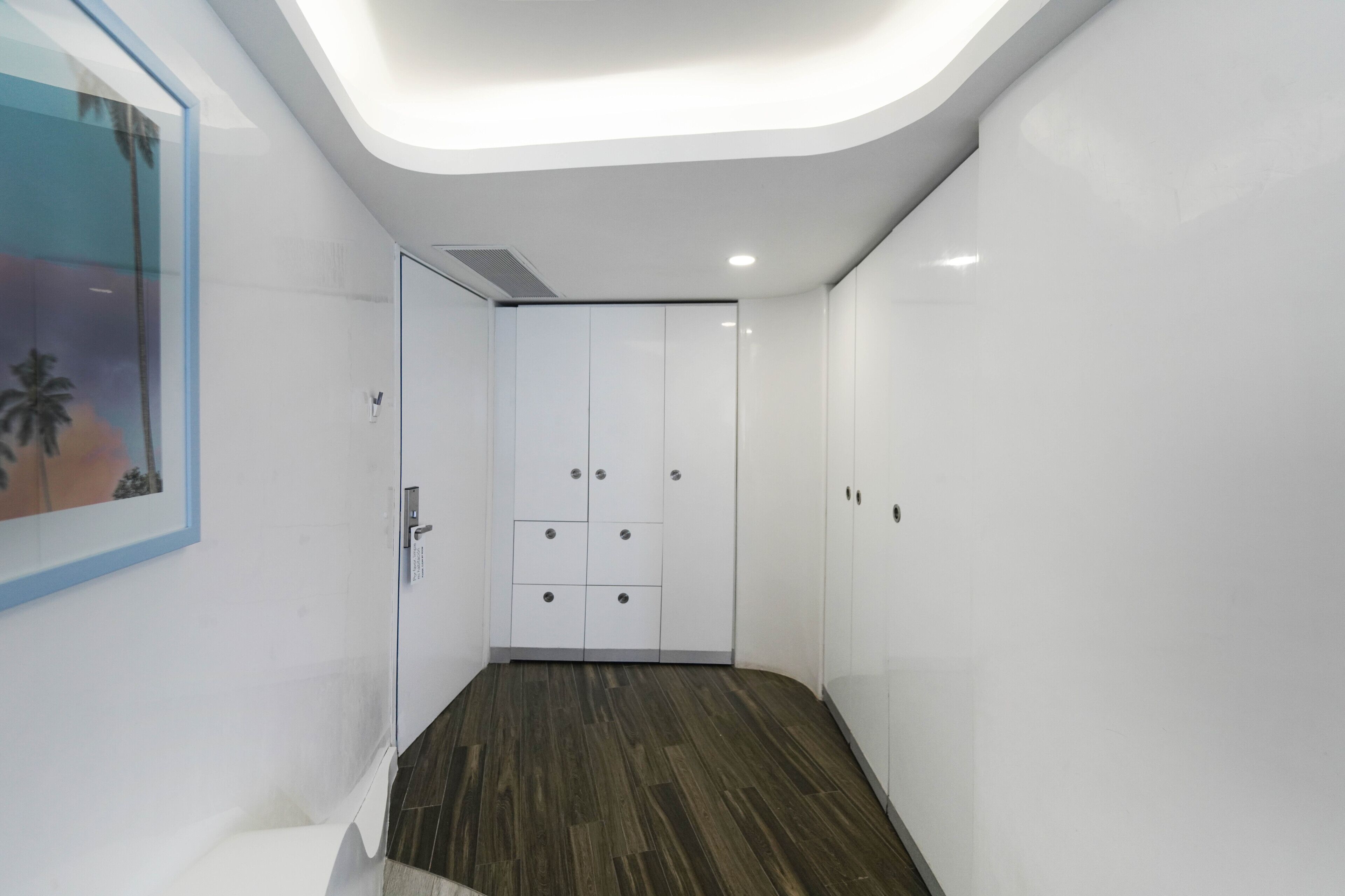 A modern hotel corridor with white walls, a wooden floor, and a large wardrobe with multiple drawers.