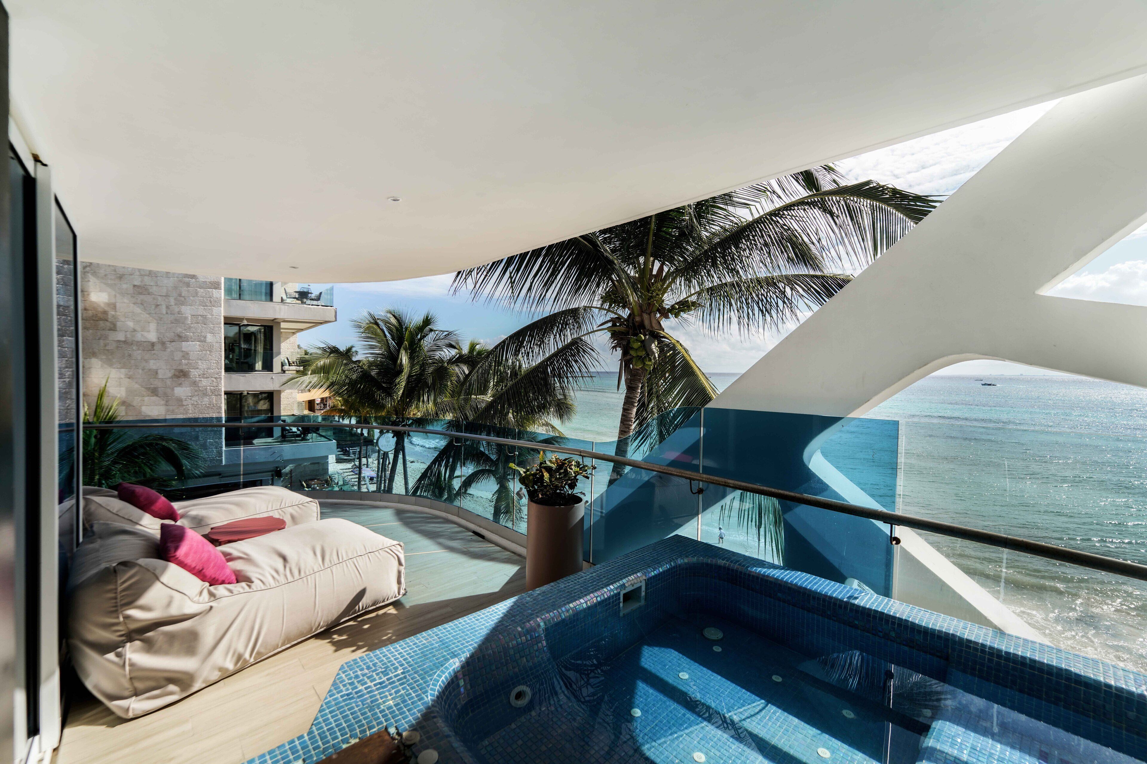 A modern balcony with a glass railing, a hot tub, and a seating area with cushions.