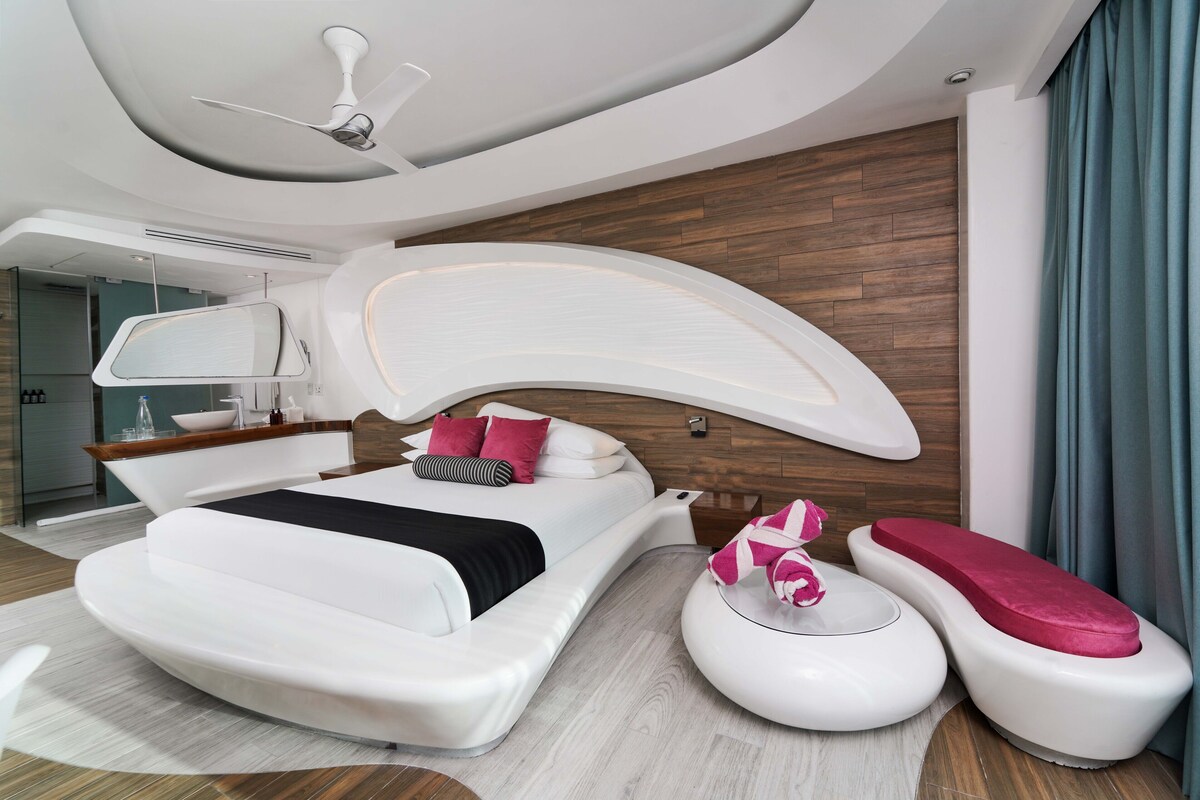 Une chambre moderne avec un plafond incurvé, un grand lit avec une literie blanche et des oreillers roses, une table de chevet et un banc intégré avec un coussin rose.
