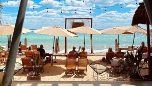 Une terrasse au bord de la plage, avec des chaises en bois et des parasols, d’où l’on a une vue sur l’océan.