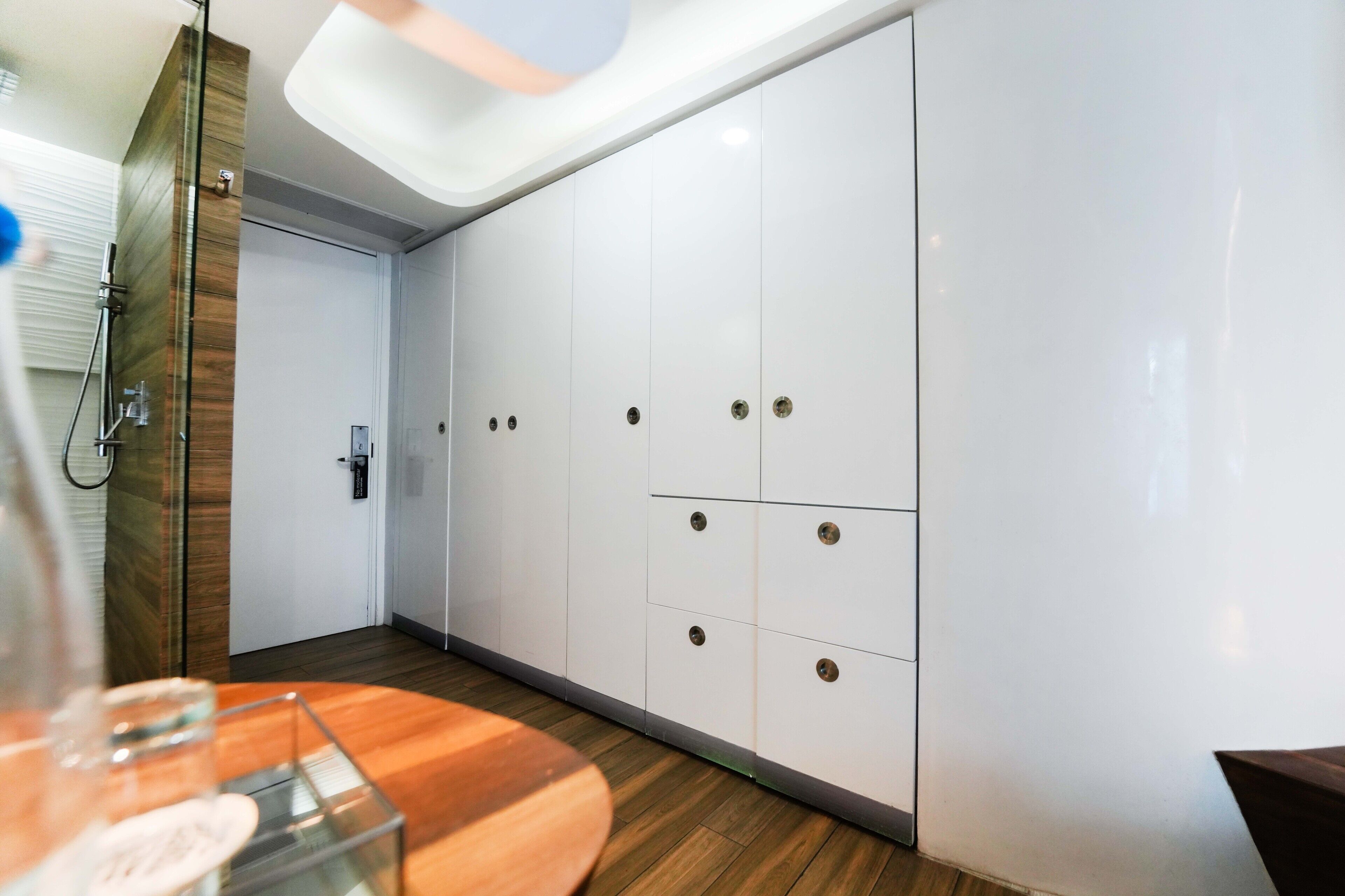 A modern hotel room with a wooden floor, white wardrobe, and a glass table.