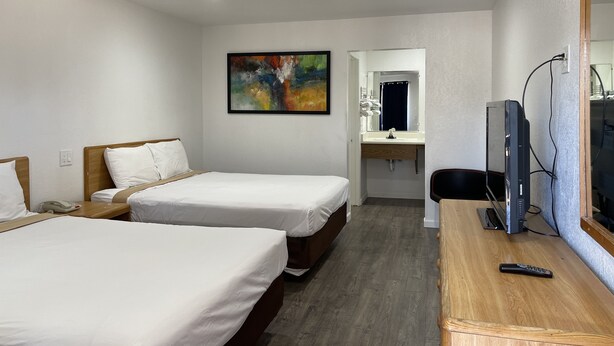 A hotel room with two beds, a desk with a TV, a bathroom mirror, and a painting on the wall. - Palms Inn & Suites (Palmdale)